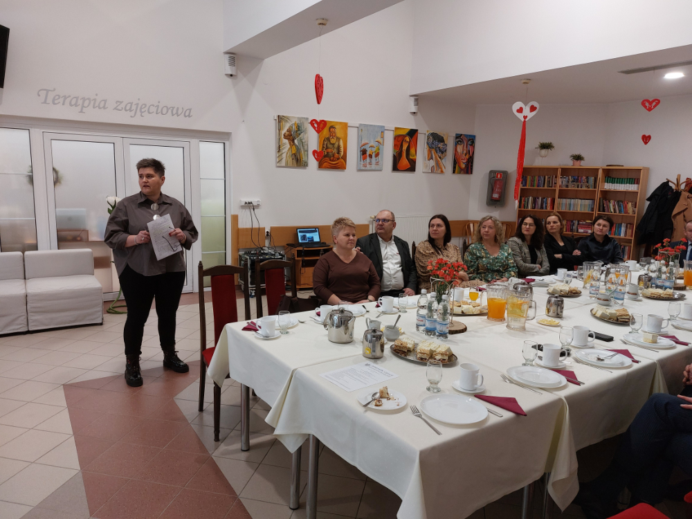 Zdjęcie przedstawia wnętrze Domu Pomocy Społecznej w Baszkowie Grupa osób na corocznym spotkaniu Zarządu Powiatu Krotoszyńskiego w osobach Starosty Krotoszyńskiego Stanisława Szczotki, Wice – Starosty Krotoszyńskiego Pawła Radojewskiego, Członka Zarządu – Miłosza Zwierzyka, Pani Sekretarz Powiatu – Joanny Dymarskiej – Kaczmarek z kadrą kierowniczą pomocy i integracji społecznej powiatu krotoszyńskiego Dyrektorzy PCPR, DPS w Baszkowie, DPS w Zdunach, ŚDS, WTZ przedstawili najważniejsze założenia w zakresie inwestycji i potrzeb wsparcia społecznego Wystąpienie Renaty Waleńskiej Dyrektor DPS w Baszkowie Pomieszczenie jest ozdobione dziełami sztuki na ścianach oraz małymi dekoracjami w kształcie serca wiszącymi z sufitu. Tekst na ścianie: Na ścianie w tle widnieje napis “Terapia zajęciowa”.