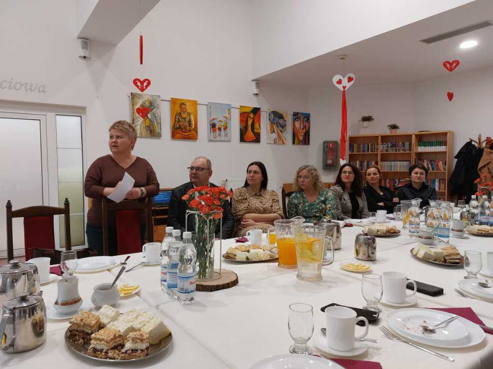 Zdjęcie przedstawia wnętrze Domu Pomocy Społecznej w Baszkowie Grupa osób na corocznym spotkaniu Zarządu Powiatu Krotoszyńskiego w osobach Starosty Krotoszyńskiego Stanisława Szczotki, Wice – Starosty Krotoszyńskiego Pawła Radojewskiego, Członka Zarządu – Miłosza Zwierzyka, Pani Sekretarz Powiatu – Joanny Dymarskiej – Kaczmarek z kadrą kierowniczą pomocy i integracji społecznej powiatu krotoszyńskiego Dyrektorzy PCPR, DPS w Baszkowie, DPS w Zdunach, ŚDS, WTZ przedstawili najważniejsze założenia w zakresie inwestycji i potrzeb wsparcia społecznego Wystąpienie Małgorzaty Nyczki Dyrektor ŚDS w Krotoszynie Pomieszczenie jest ozdobione dziełami sztuki na ścianach oraz małymi dekoracjami w kształcie serca wiszącymi z sufitu. Tekst na ścianie: Na ścianie w tle widnieje napis “Terapia zajęciowa”.