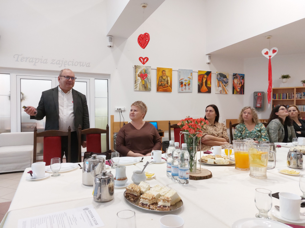 Zdjęcie przedstawia wnętrze Domu Pomocy Społecznej w Baszkowie Grupa osób na corocznym spotkaniu Zarządu Powiatu Krotoszyńskiego w osobach Starosty Krotoszyńskiego Stanisława Szczotki, Wice – Starosty Krotoszyńskiego Pawła Radojewskiego, Członka Zarządu – Miłosza Zwierzyka, Pani Sekretarz Powiatu – Joanny Dymarskiej – Kaczmarek z kadrą kierowniczą pomocy i integracji społecznej powiatu krotoszyńskiego Dyrektorzy PCPR, DPS w Baszkowie, DPS w Zdunach, ŚDS, WTZ przedstawili najważniejsze założenia w zakresie inwestycji i potrzeb wsparcia społecznego Wystąpienie Dyrektora WTZ w Krotoszynie Pomieszczenie jest ozdobione dziełami sztuki na ścianach oraz małymi dekoracjami w kształcie serca wiszącymi z sufitu. Tekst na ścianie: Na ścianie w tle widnieje napis “Terapia zajęciowa”.