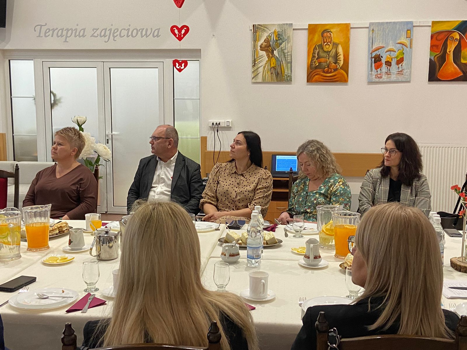 Zdjęcie przedstawia wnętrze Domu Pomocy Społecznej w Baszkowie Grupa osób na corocznym spotkaniu Zarządu Powiatu Krotoszyńskiego w osobach Starosty Krotoszyńskiego Stanisława Szczotki, Wice – Starosty Krotoszyńskiego Pawła Radojewskiego, Członka Zarządu – Miłosza Zwierzyka, Pani Sekretarz Powiatu – Joanny Dymarskiej – Kaczmarek z kadrą kierowniczą pomocy i integracji społecznej powiatu krotoszyńskiego Dyrektorzy PCPR, DPS w Baszkowie, DPS w Zdunach, ŚDS, WTZ przedstawili najważniejsze założenia w zakresie inwestycji i potrzeb wsparcia społecznego Pomieszczenie jest ozdobione dziełami sztuki na ścianach oraz małymi dekoracjami w kształcie serca wiszącymi z sufitu. Tekst na ścianie: Na ścianie w tle widnieje napis “Terapia zajęciowa”.