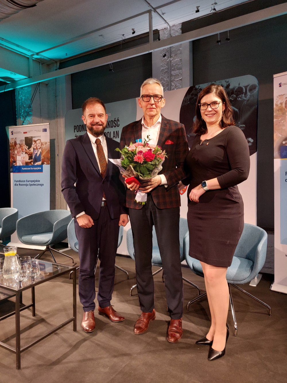 Kobieta i dwóch mężczyzn w garniturach, Dyrektor PCPR trzyma w ręku wiązankę kwiatów