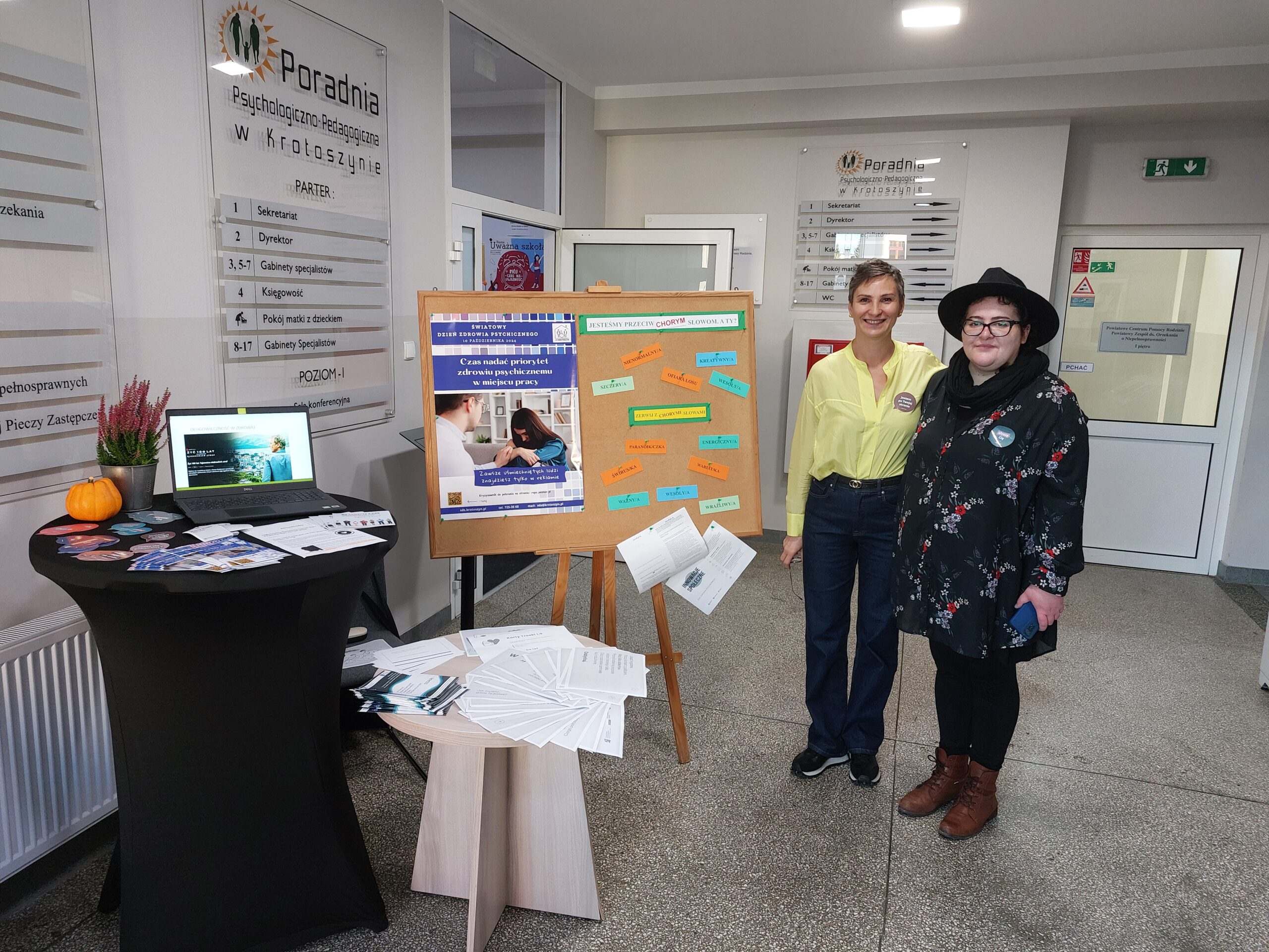 Wystawa w pomieszczeniu, tablica z plakatem i kartkami z napisami, na stoliku komputer i ulotki.
Dwie kobiety prowadzące wystawę