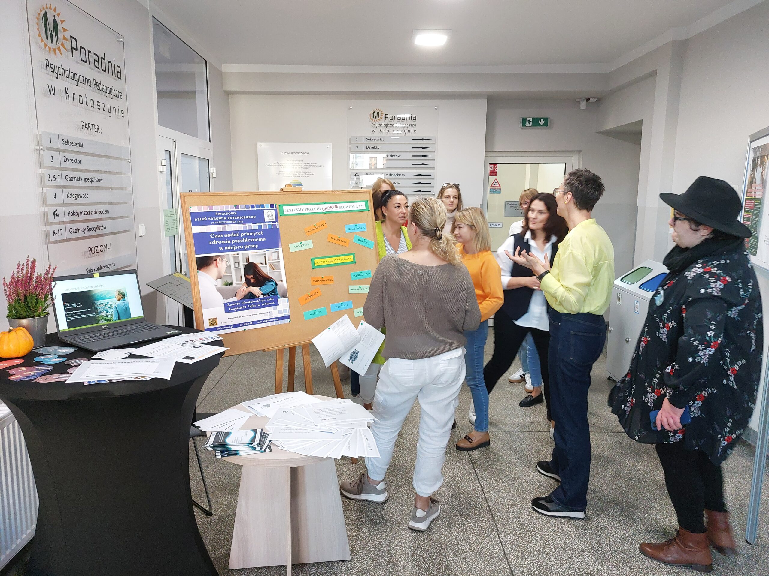 Wystawa w pomieszczeniu, tablica z plakatem i kartkami z napisami, na stoliku komputer i ulotki.
Dwie kobiety prowadzące wystawę oraz osoby wystawę oglądające