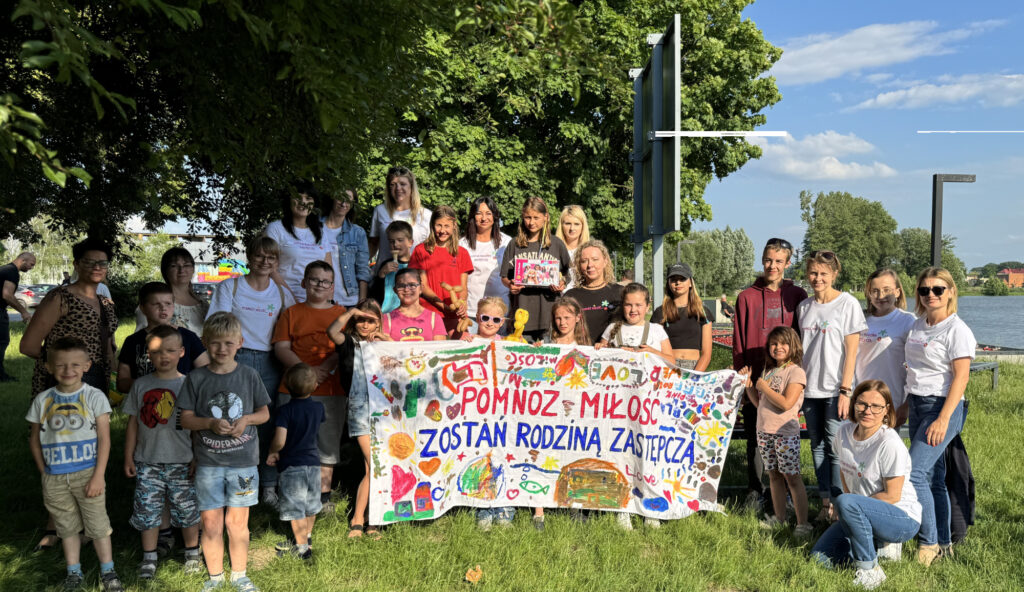 Grupa ludzi, w tym dorośli i dzieci, stoi na zewnątrz na trawiastym terenie w pobliżu zbiornika wodnego. Trzymają kolorowy baner z różnymi rysunkami i tekstem w języku polskim. Na banerze widnieje napis: 'POMNOŻ MIŁOŚĆ ZOSTAŃ RODZINĄ ZASTĘPCZĄ.' Scena jest wydarzeniem promującym opiekę zastępczą. W tle widać drzewa, czyste niebo oraz kilka odległych budynków.