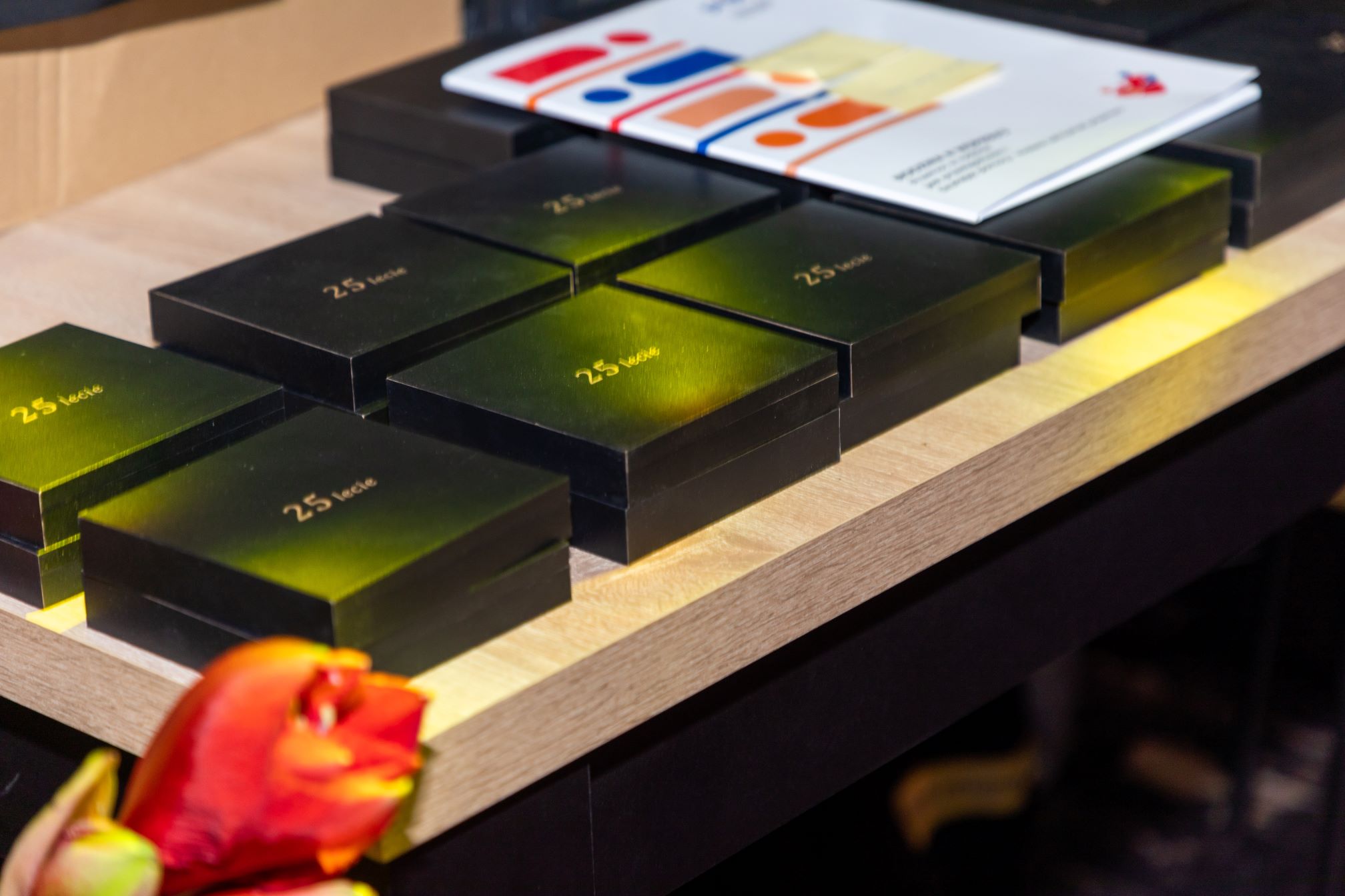 Na zdjęciu widoczne są czarne pudełka z napisem '25 lecie' ułożone na drewnianym blacie. W tle znajduje się broszura z kolorowymi grafikami. Na pierwszym planie widoczny jest czerwony kwiat.
