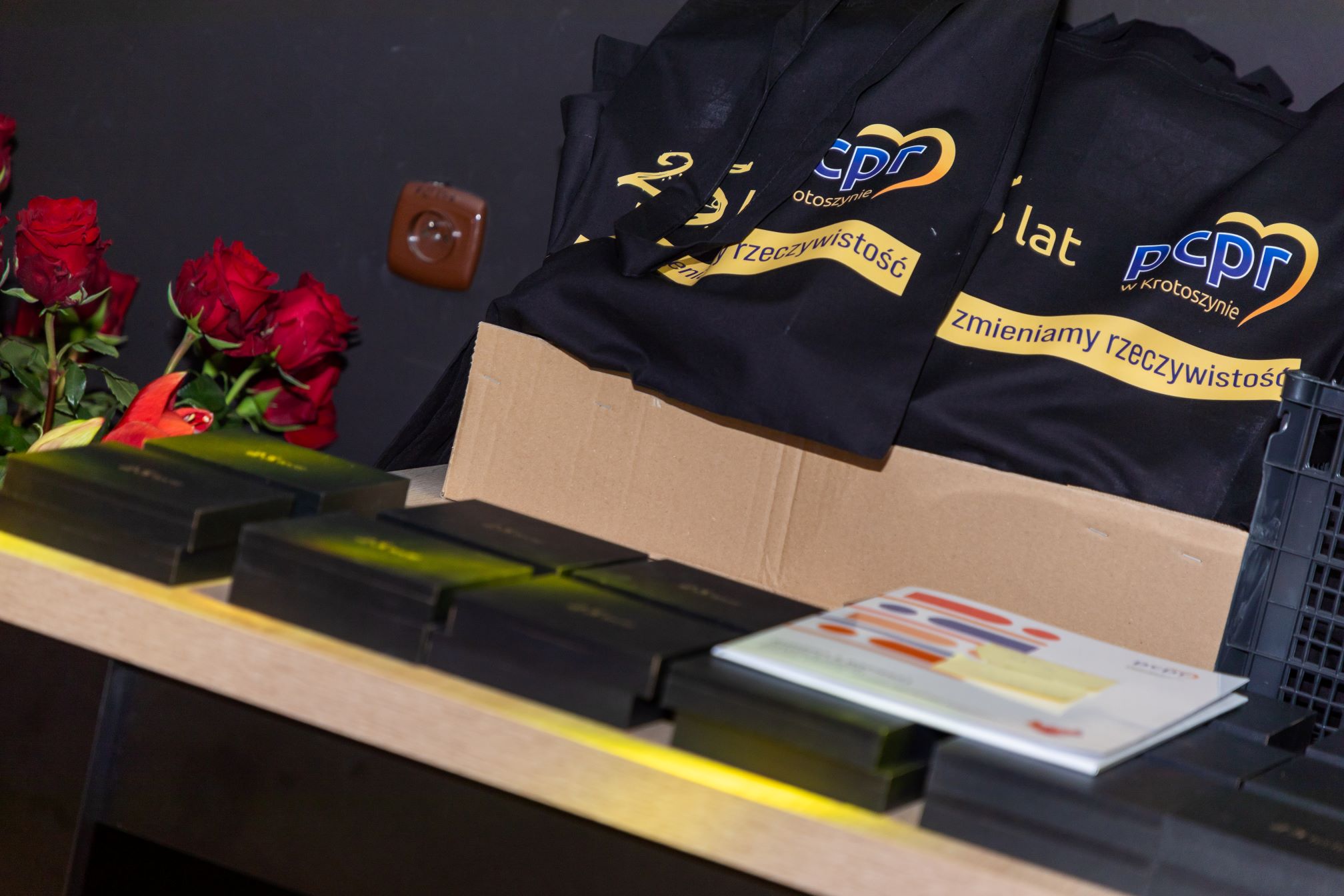 Na zdjęciu widoczne są czarne torby z logo PCPR oraz napisem 'zmieniamy rzeczywistość' i 'w Krotoszynie'. Torby znajdują się na kartonowym pudełku, a obok nich leżą czerwone róże oraz kilka czarnych pudełek. Na stole znajduje się również broszura lub katalog z kolorowymi grafikami.
