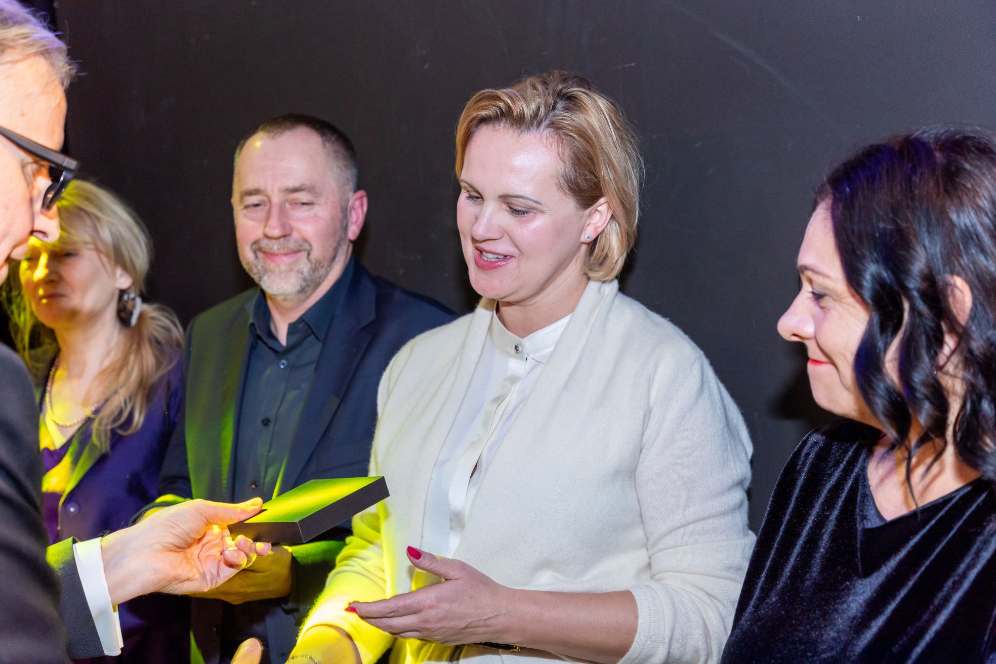 Na zdjęciu znajduje się grupa osób stojących w rzędzie. Osoby te są ubrane elegancko. Jedna z osób, znajdująca się po lewej stronie, trzyma w rękach prostokątny przedmiot, który może być dyplomem, certyfikatem lub nagrodą. Tło jest ciemne, co sprawia, że osoby na pierwszym planie są dobrze widoczne.
