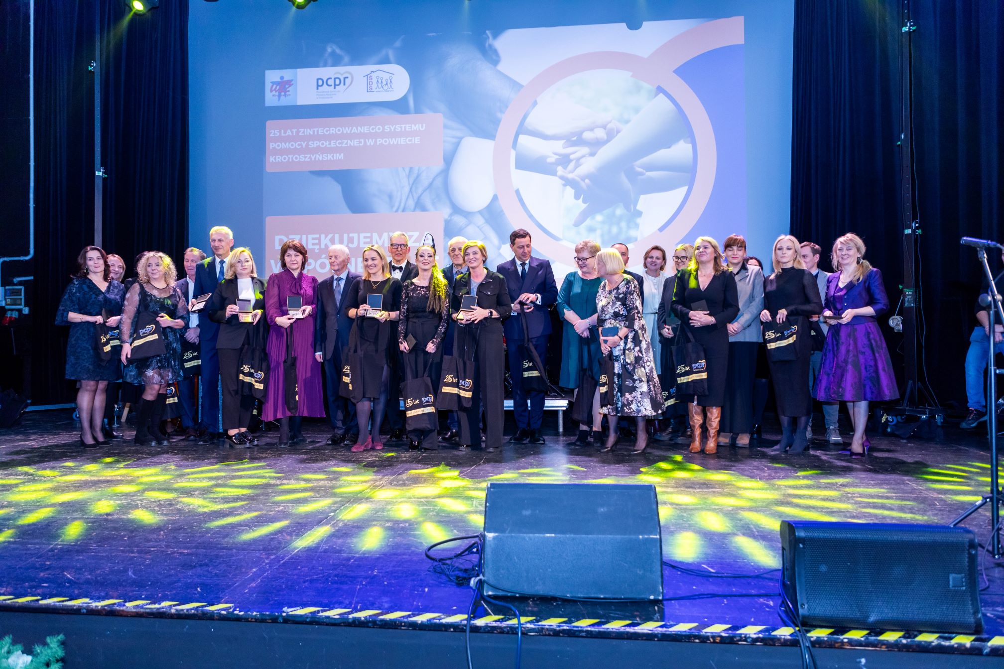 Na zdjęciu znajduje się grupa ludzi stojących na scenie podczas uroczystości. W tle widoczny jest ekran z napisem: "25 LAT ZINTEGROWANEGO SYSTEMU POMOCY SPOŁECZNEJ W POWIECIE KROTOSZYŃSKIM" oraz "DZIĘKUJEMY ZA WSPÓŁPRACĘ". Na ekranie znajdują się również logotypy PCPR oraz innych organizacji. Osoby na scenie trzymają w rękach nagrody i torby z napisem "25 lat". Scena jest oświetlona kolorowymi światłami, a w tle widać elementy dekoracyjne.
