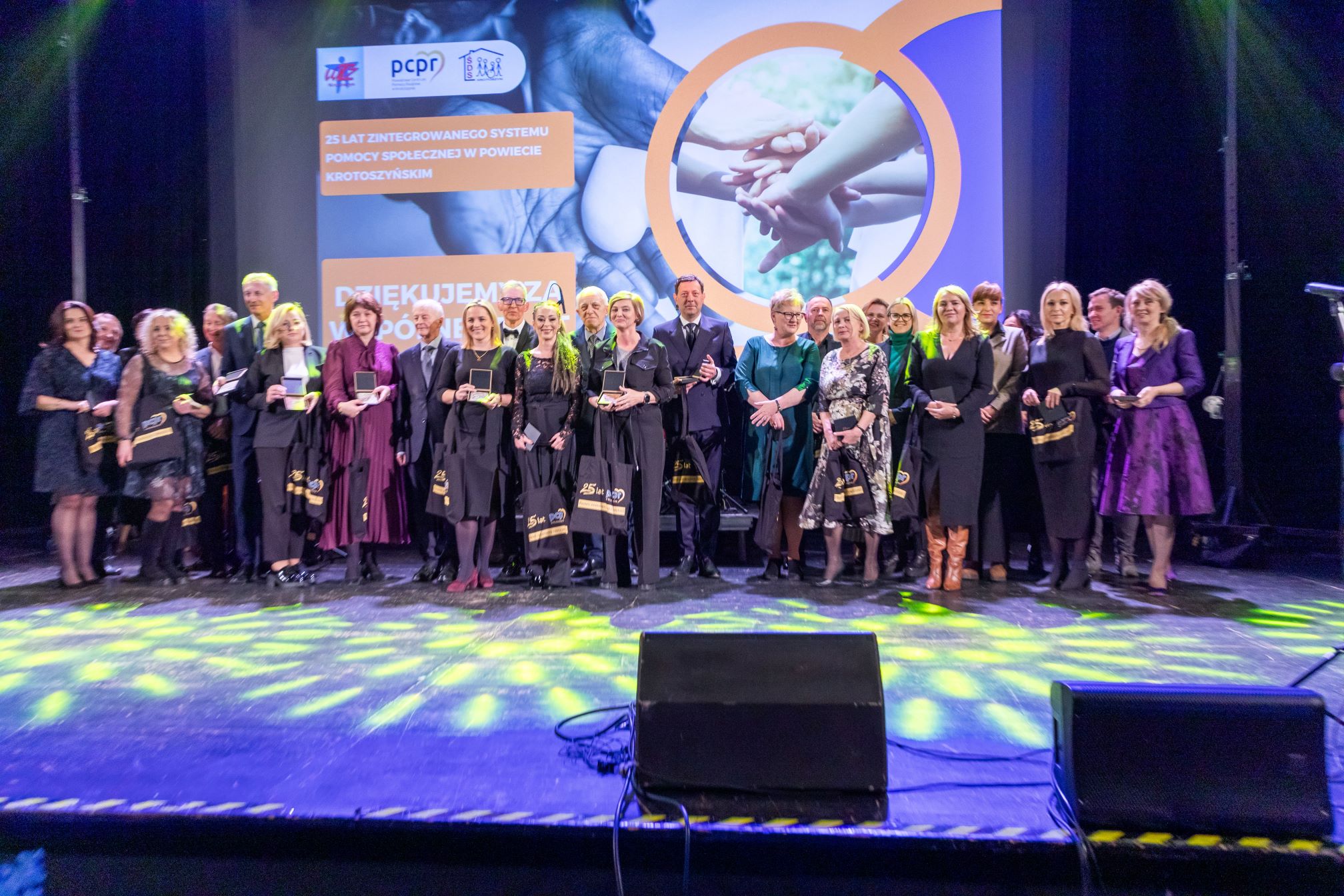 Na zdjęciu znajduje się grupa ludzi stojących na scenie. W tle widoczny jest ekran z napisem: '25 LAT ZINTEGROWANEGO SYSTEMU POMOCY SPOŁECZNEJ W POWIECIE KROTOSZYŃSKIM'. Na ekranie znajdują się również logotypy organizacji oraz zdjęcie przedstawiające złączone ręce. Osoby na scenie trzymają w rękach nagrody i torby z napisem '25 lat'. Scena jest oświetlona kolorowymi światłami, co nadaje uroczysty charakter wydarzeniu
