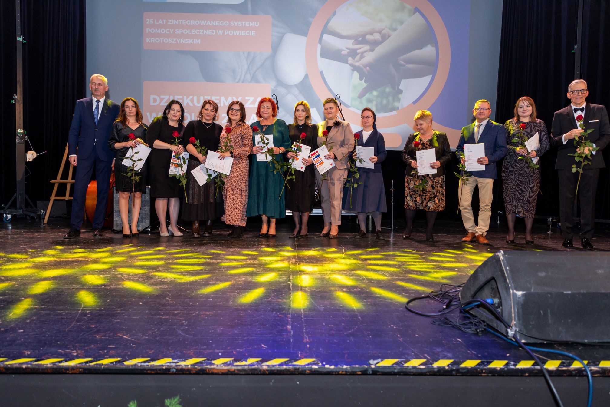 Na zdjęciu znajduje się grupa ludzi stojących na scenie. Każda osoba trzyma w ręku różę oraz dokumenty, prawdopodobnie certyfikaty lub dyplomy. W tle widoczny jest ekran z napisem: '25 LAT ZINTEGROWANEGO SYSTEMU POMOCY SPOŁECZNEJ W POWIECIE KROTOSZYŃSKIM' oraz 'DZIĘKUJEMY ZA WSPÓŁPRACĘ'. Na ekranie znajduje się również obrazek przedstawiający splecione ręce. Scena jest oświetlona żółtymi reflektorami, a na podłodze widać ich odbicia.
