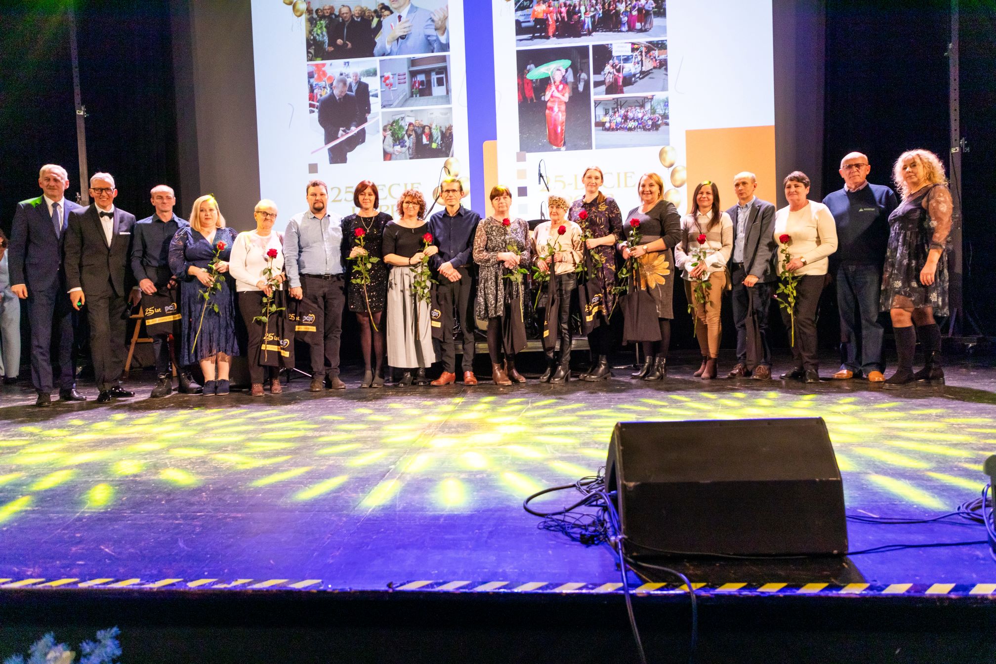 Zdjęcie przedstawia grupę ludzi stojących na scenie, trzymających kwiaty i torby z prezentami. Wydarzenie wygląda na formalne, prawdopodobnie rocznicę lub ceremonię wręczenia nagród. W tle znajduje się duży ekran wyświetlający różne zdjęcia i tekst, w tym numer '25' i słowo 'LECIE', co sugeruje, że jest to obchody 25-lecia. Scena jest oświetlona kolorowymi światłami, co dodaje atmosfery uroczystości. Osoby są ubrane w stroje formalne lub półformalne, co podkreśla znaczenie tego wydarzenia.
