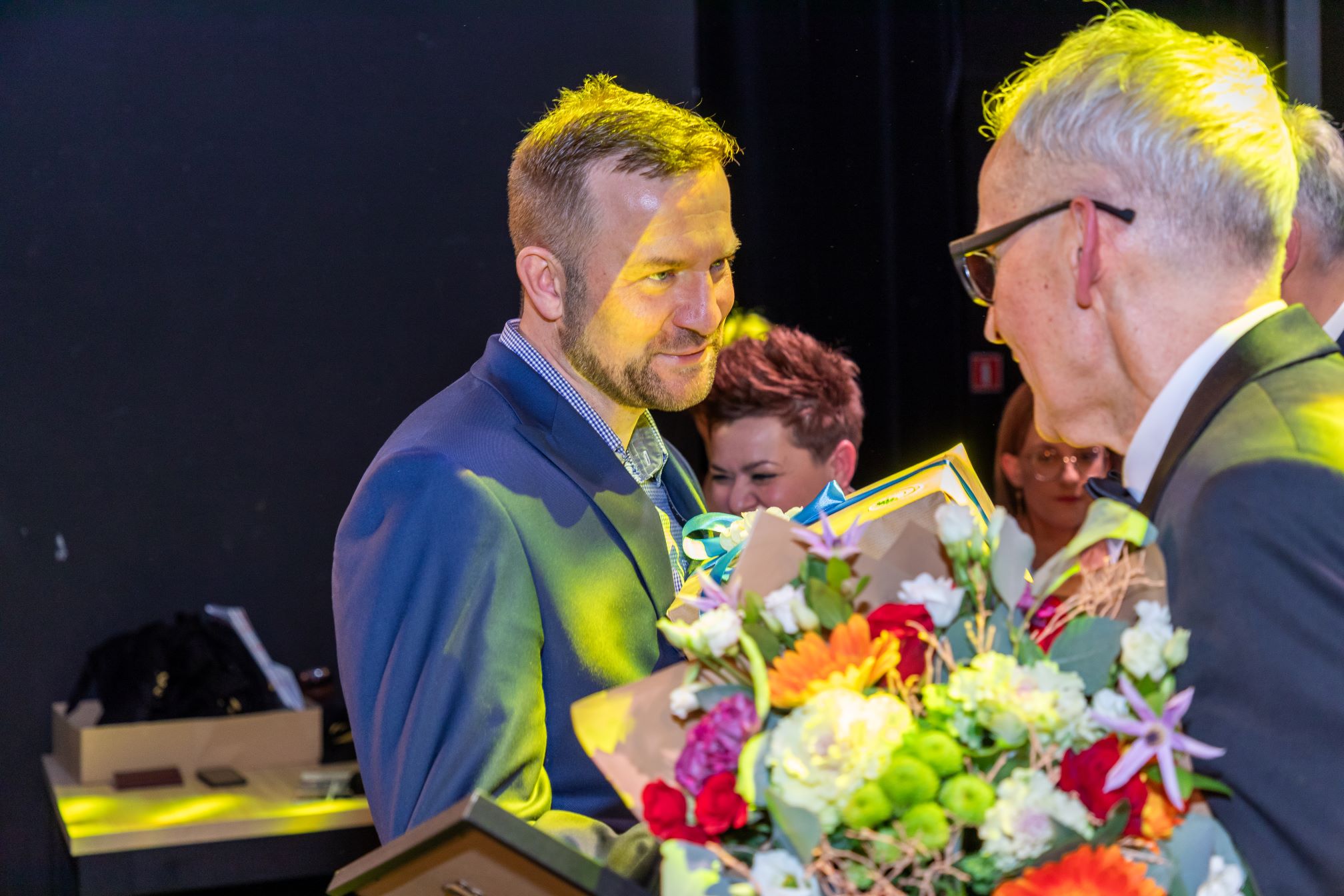 Na zdjęciu widać dwie osoby w eleganckich strojach, które prawdopodobnie uczestniczą w ceremonii wręczenia nagród. Jedna z osób trzyma bukiet kwiatów, a druga trzyma dyplom lub certyfikat. W tle widać inne osoby oraz stół z różnymi przedmiotami.
