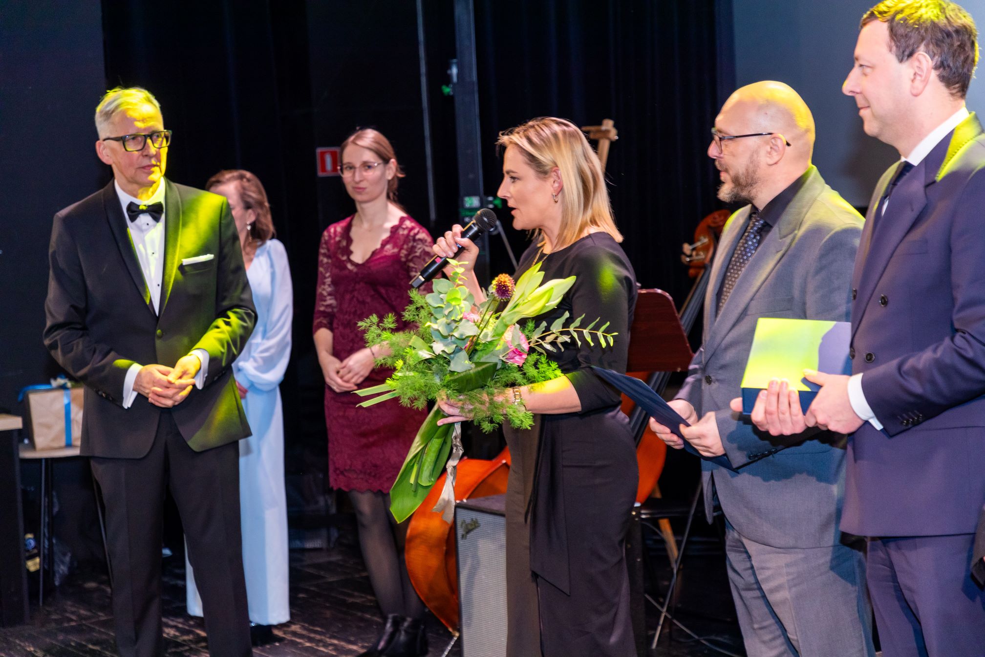 Na zdjęciu znajduje się grupa osób na scenie podczas uroczystości. Jedna z osób trzyma mikrofon i bukiet kwiatów, prawdopodobnie przemawia. Inne osoby stoją obok, trzymając dokumenty lub dyplomy. W tle widać instrumenty muzyczne.
