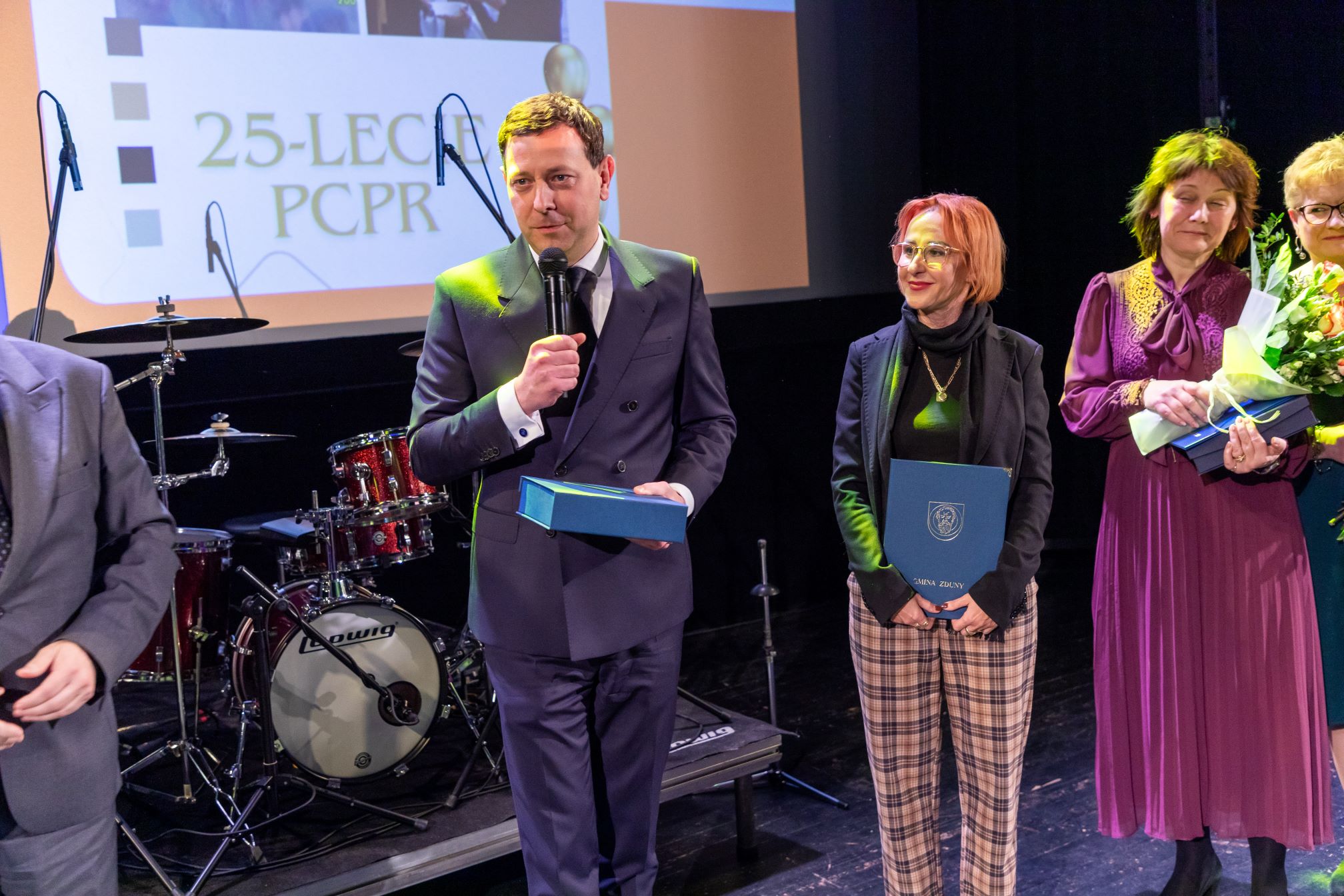 Na zdjęciu znajduje się grupa osób stojących na scenie podczas uroczystości. W tle widać perkusję oraz ekran z napisem '25-LECIE PCPR'. Osoba po lewej stronie trzyma mikrofon i niebieskie pudełko, a obok niej stoi kobieta trzymająca niebieską teczkę z napisem 'GMINA ZDUNY'. Po prawej stronie znajduje się kobieta w fioletowej sukience, trzymająca bukiet kwiatów. Cała scena jest oświetlona, co sugeruje, że jest to oficjalne wydarzenie lub ceremonia wręczenia nagród.
