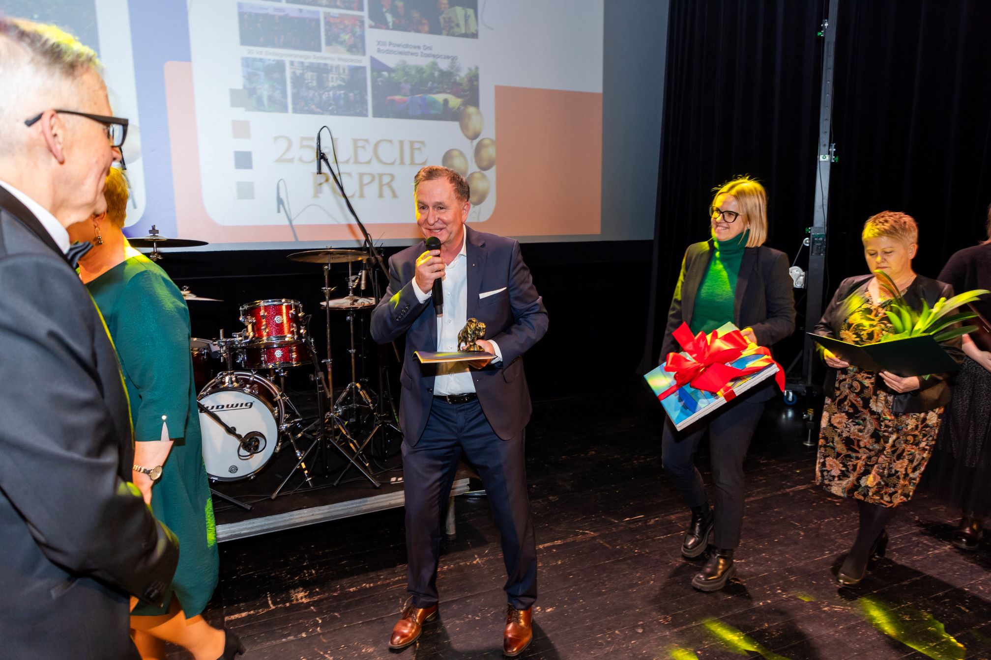 Na zdjęciu znajduje się grupa osób na scenie podczas uroczystości. W tle widać ekran z napisem '25 LECIE PCPR'. Na scenie znajduje się zestaw perkusyjny marki Ludwig. Jedna osoba trzyma mikrofon i statuetkę, a dwie inne osoby trzymają prezenty i kwiaty.

