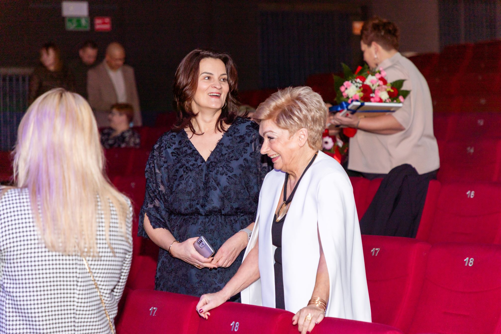 Na zdjęciu widać kilka osób w sali kinowej. Dwie osoby stoją przy rzędzie czerwonych foteli oznaczonych numerami 17 i 18. Jedna z nich trzyma telefon komórkowy. W tle widać inne osoby, w tym jedną trzymającą bukiet kwiatów.
