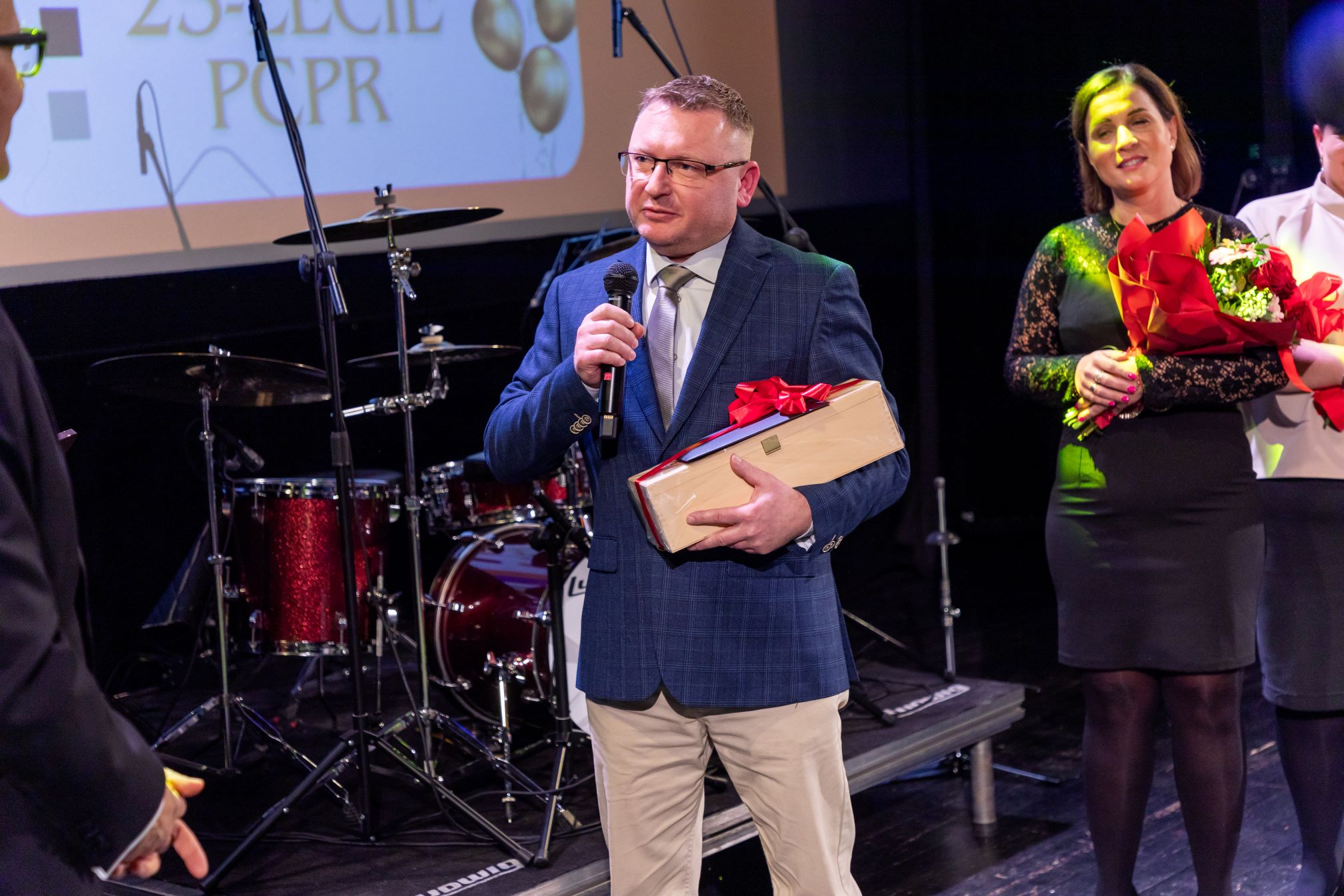 Na zdjęciu znajduje się mężczyzna w garniturze, trzymający mikrofon i zapakowany prezent z czerwoną wstążką. Obok niego stoi kobieta trzymająca bukiet kwiatów. W tle widać zestaw perkusyjny oraz ekran z napisem '25-LECIE PCPR'. Scena sugeruje, że jest to uroczystość związana z jubileuszem 25-lecia.
