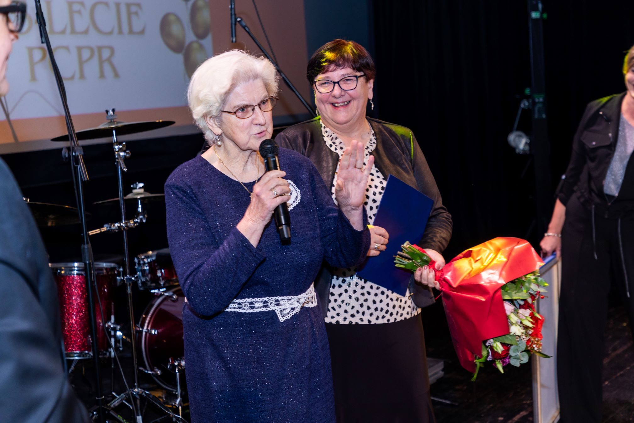 Na zdjęciu znajduje się kobieta trzymająca mikrofon i przemawiająca do publiczności. Obok niej stoi inna kobieta trzymająca bukiet kwiatów owinięty w czerwony papier, trzyma niebieską teczkę. W tle widać zestaw perkusyjny oraz fragment ekranu z napisem 'LECIE PCPR'. Zdjęcie zostało wykonane podczas jakiegoś wydarzenia lub uroczystości, co sugeruje obecność mikrofonu, kwiatów oraz formalnego ubioru osób na zdjęciu.
