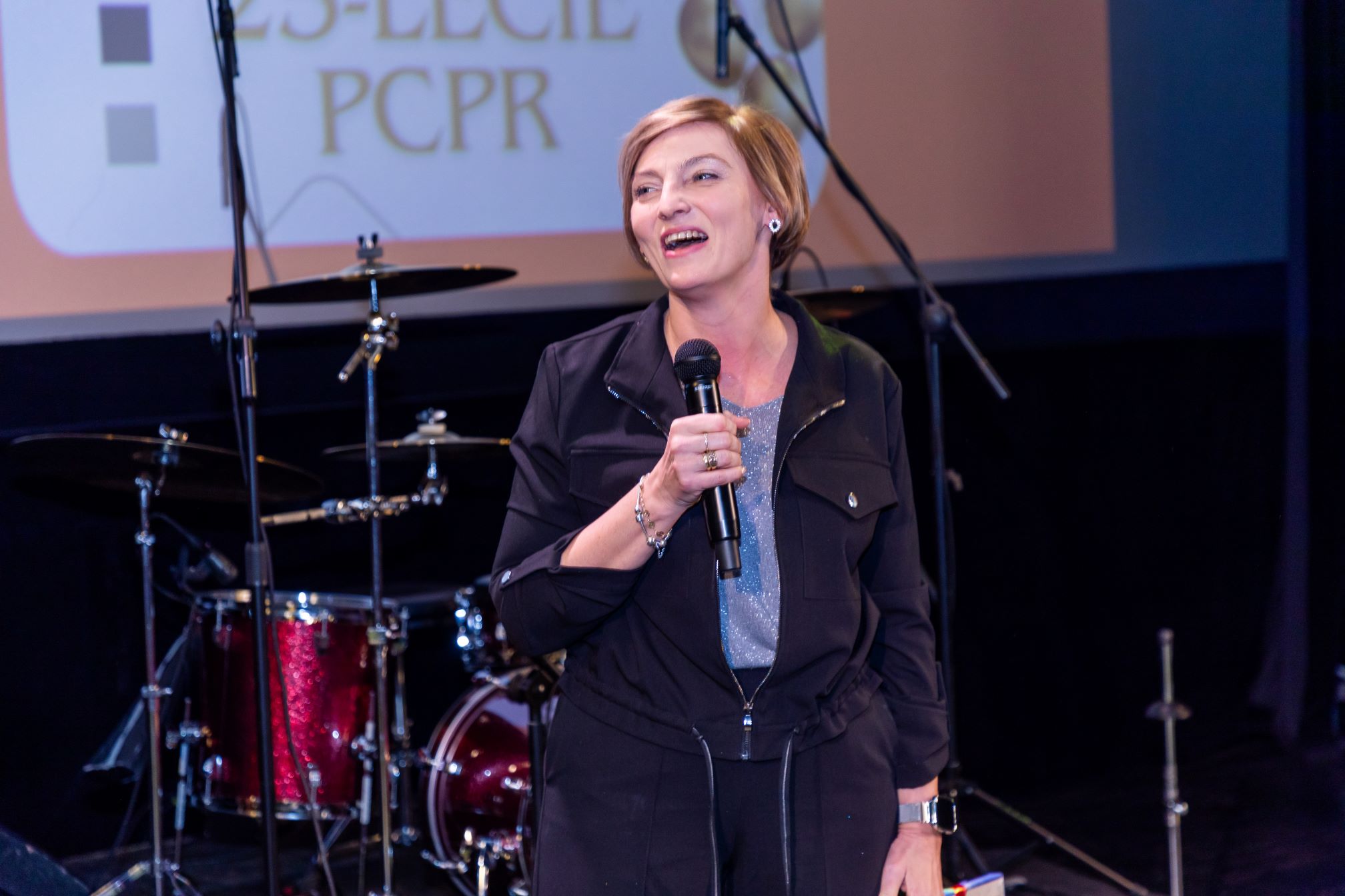 Na zdjęciu znajduje się osoba trzymająca mikrofon, stojąca na scenie przed zestawem perkusyjnym. W tle widać ekran z napisem "25-LECIE PCPR".
