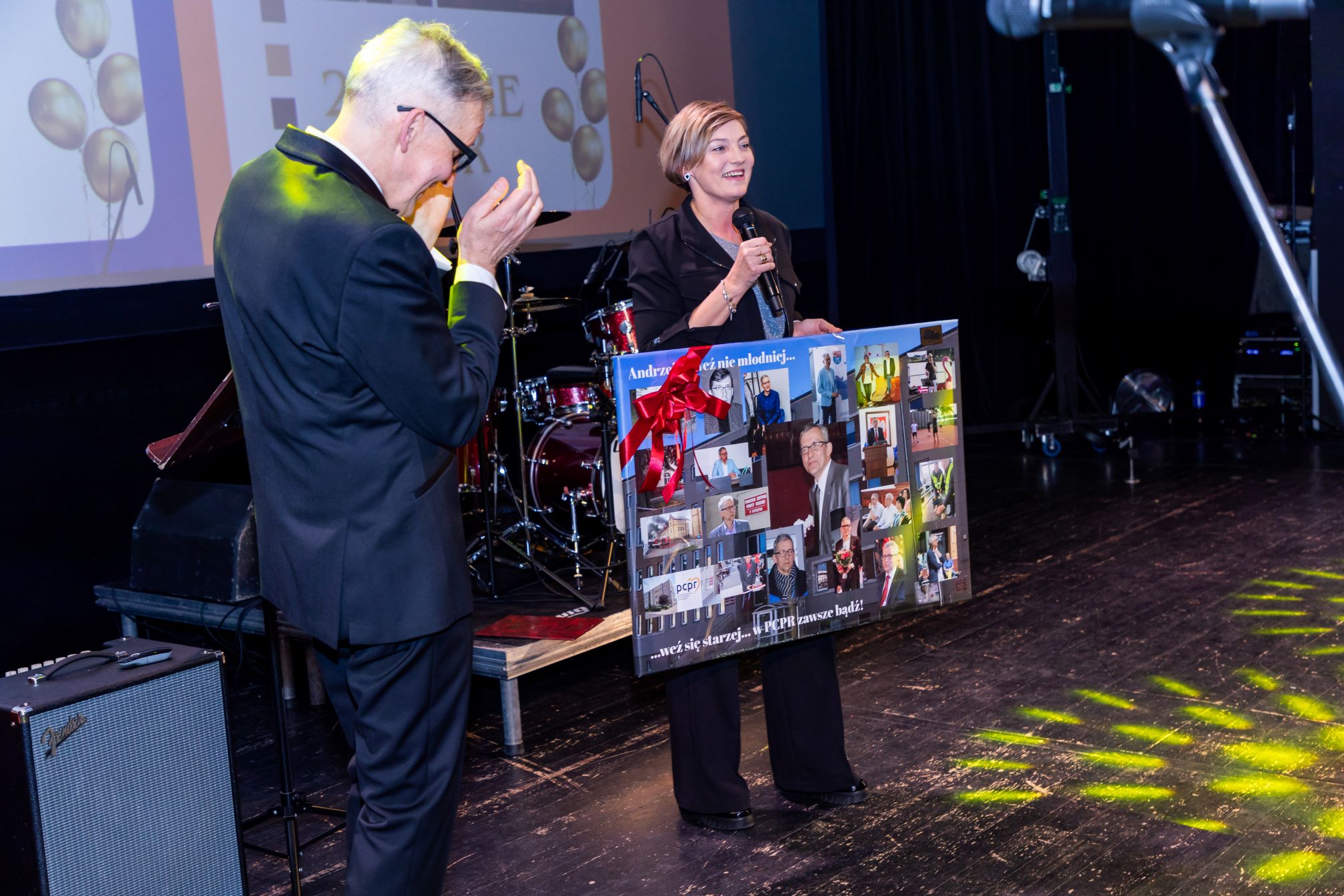 Na zdjęciu widać dwie osoby na scenie podczas uroczystości. Jedna z nich trzyma mikrofon, a druga trzyma dużą tablicę z wieloma zdjęciami i napisem 'Andrzeju też nie młodniejesz... weź się starzej... i PCPR zawsze bądź!'. W tle widać perkusję i inne elementy sceniczne.

