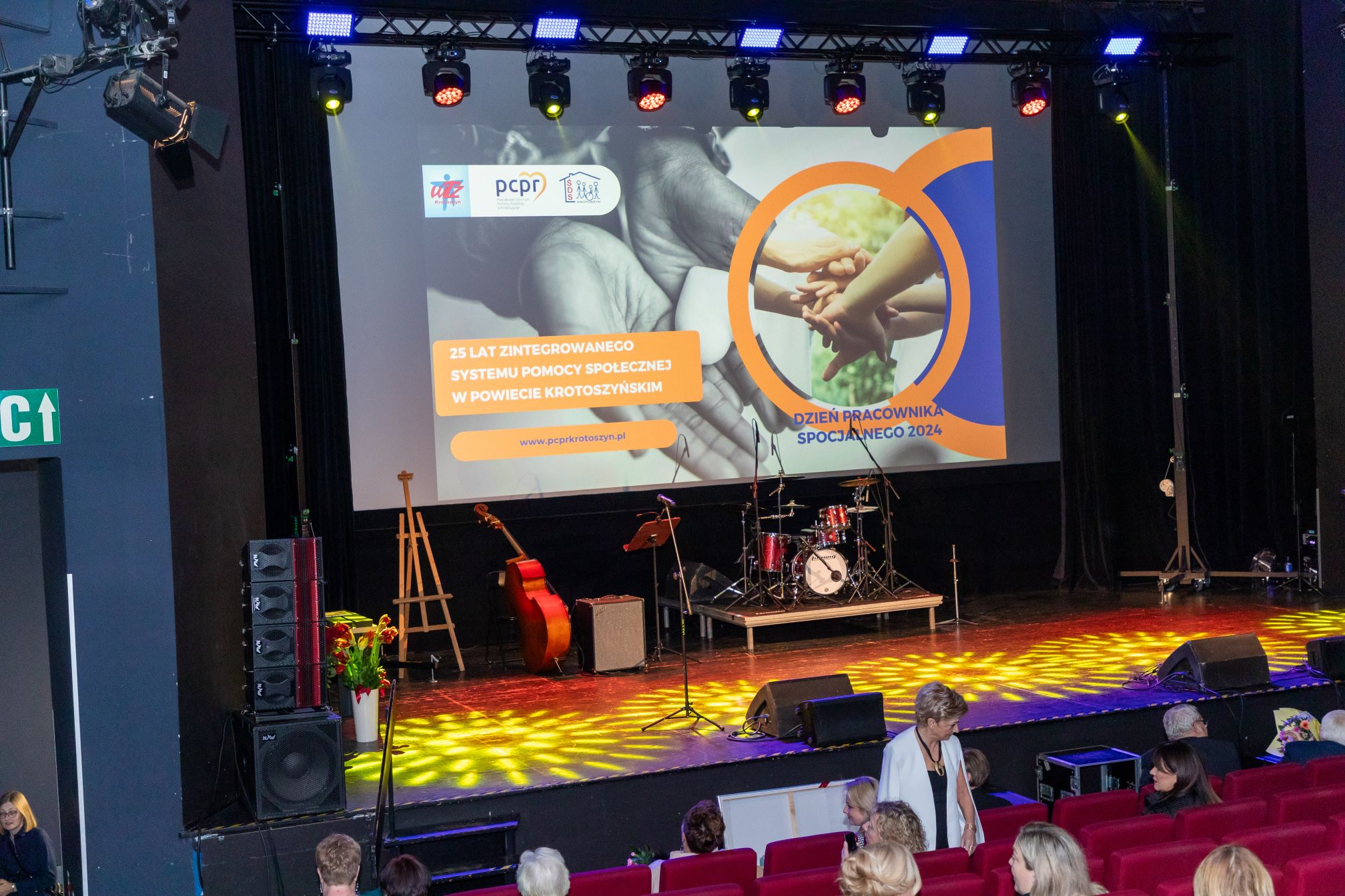 Na zdjęciu znajduje się scena w sali kinowej, na której widać instrumenty muzyczne, takie jak perkusja, kontrabas i mikrofon. Na ekranie wyświetlany jest slajd z napisem '25 LAT ZINTEGROWANEGO SYSTEMU POMOCY SPOŁECZNEJ W POWIECIE KROTOSZYŃSKIM' oraz 'DZIEŃ PRACOWNIKA SOCJALNEGO 2024'. Na slajdzie znajdują się również logotypy organizacji oraz adres strony internetowej www.pcprkrotoszyn.pl.Widzowie siedzą na czerwonych fotelach, a niektórzy z nich stoją przed sceną.
