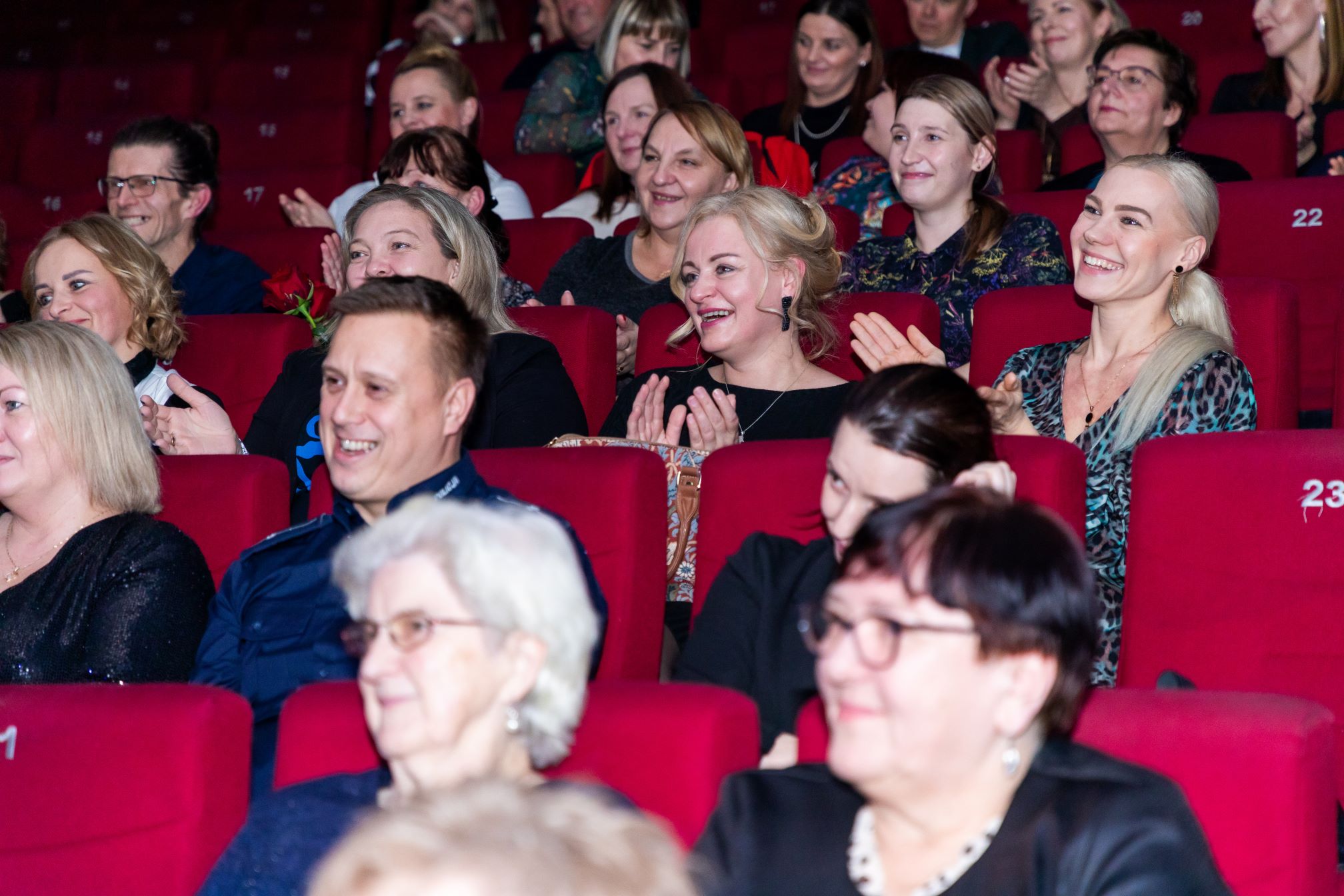 Na zdjęciu widać grupę ludzi siedzących w kinie. Wszyscy siedzą na czerwonych fotelach, a niektórzy z nich klaszczą. W tle widoczne są inne osoby, również siedzące, obserwujące coś poza kadrem. Na fotelach widoczne są numery miejsc, takie jak '22' i '23'.
