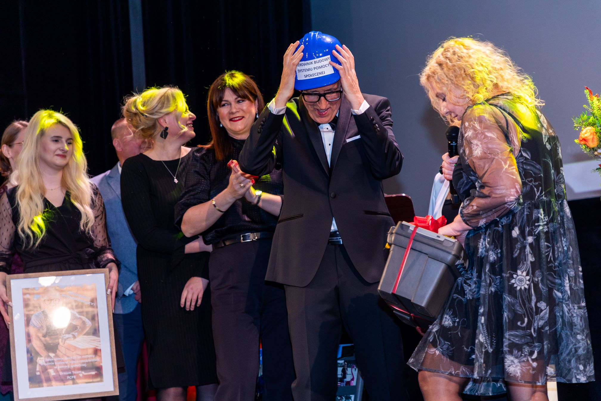 Na zdjęciu znajduje się grupa osób na scenie. Jedna z osób, ubrana w garnitur, zakłada niebieski kask ochronny z napisem 'KIEROWNIK BUDOWY SYSTEMU POMOCY SPOŁECZNEJ'. Obok tej osoby stoi kobieta w czarnej sukience, trzymająca mikrofon i pudełko z czerwoną wstążką. W tle widać inne osoby, z których jedna trzyma w rękach oprawiony obraz. Cała scena jest oświetlona reflektorami.
