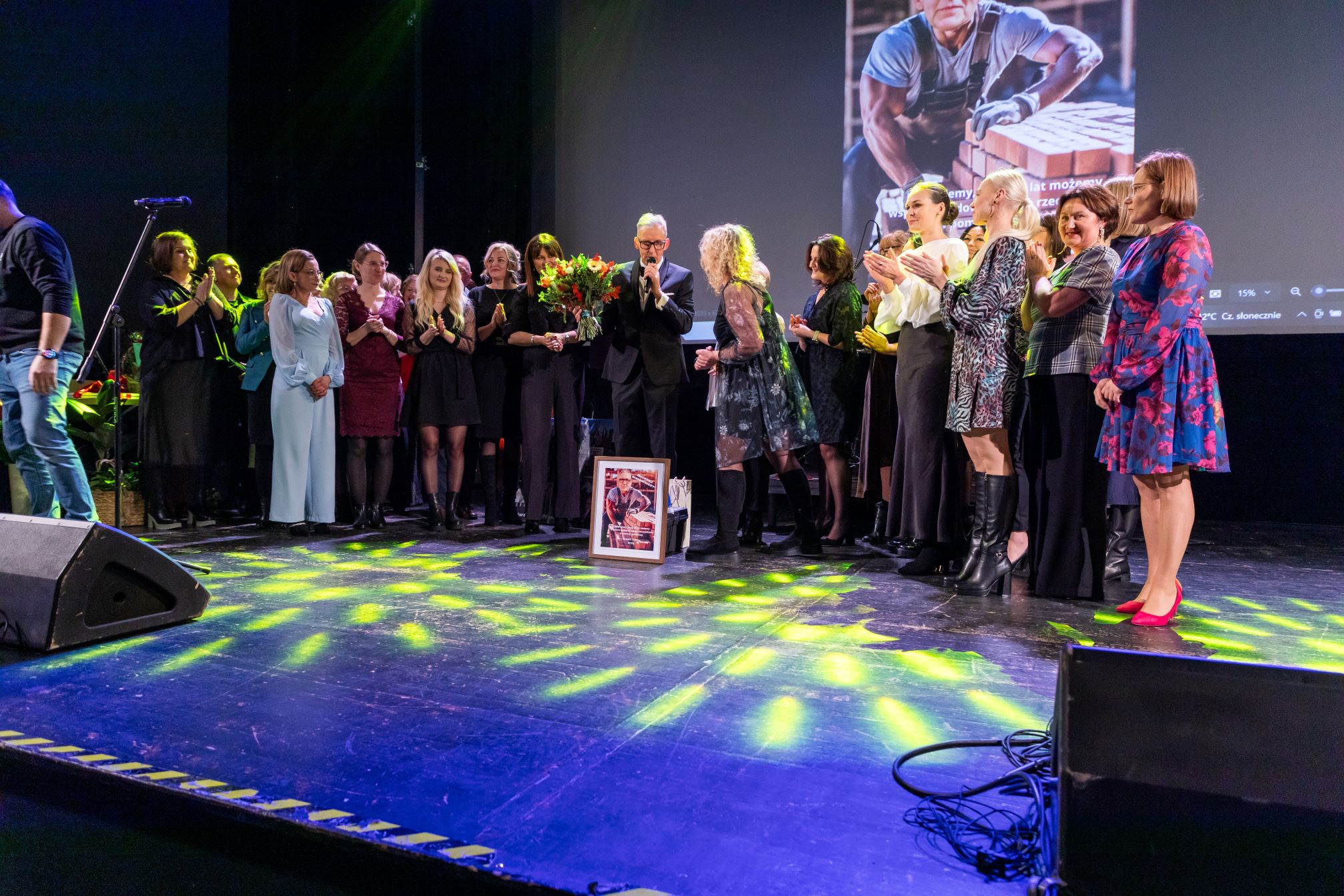 Na zdjęciu znajduje się grupa ludzi stojących na scenie. W centrum grupy znajduje się osoba trzymająca bukiet kwiatów, a przed nią na podłodze stoi oprawione zdjęcie. W tle widać duży ekran z wyświetlonym obrazem przedstawiającym osobę pracującą z cegłami. Na ekranie widnieje również tekst w języku polskim. Scena jest oświetlona kolorowymi światłami, a po lewej stronie widać fragment głośnika i mikrofonu na statywie. Wszyscy ludzie na scenie są elegancko ubrani.
