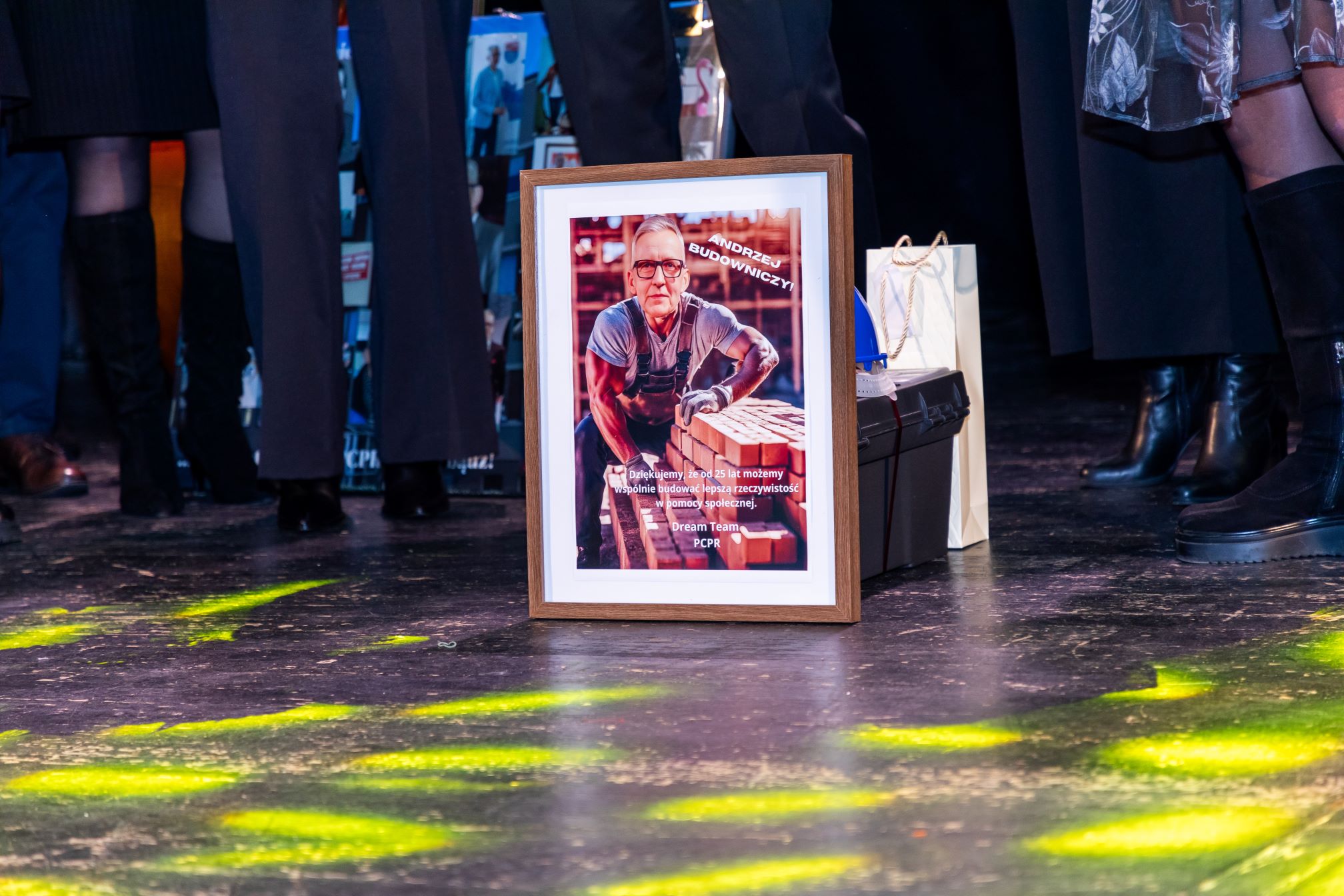 Na zdjęciu widać grupę ludzi stojących na scenie. Na pierwszym planie znajduje się oprawione w ramkę zdjęcie mężczyzny pracującego na budowie. Na zdjęciu w ramce widnieje napis: 'ANDRZEJ BUDOWNICZY'. Poniżej znajduje się tekst: 'Dziękujemy, że od 25 lat możemy wspólnie budować lepszą rzeczywistość pomocy społecznej. Dream Team PCPR'. Obok ramki znajdują się torby z prezentami.
