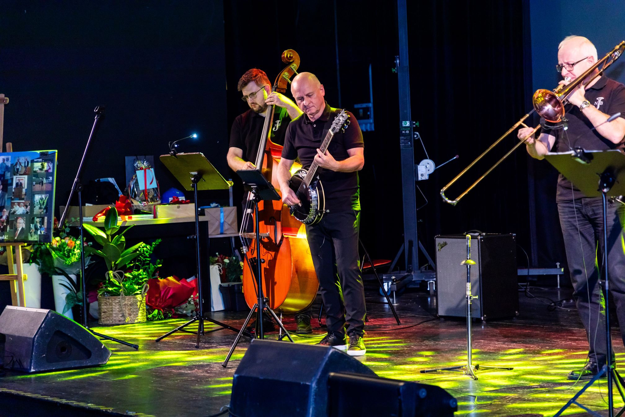 Na zdjęciu znajduje się scena, na której występuje zespół muzyczny. W skład zespołu wchodzą trzy osoby grające na różnych instrumentach: kontrabasie, banjo i puzonie. W tle widać różne dekoracje, w tym rośliny doniczkowe i tablicę ze zdjęciami. Na scenie znajdują się również mikrofony i stojaki na nuty. Cała scena jest oświetlona kolorowymi światłami, co dodaje atmosfery występowi.
