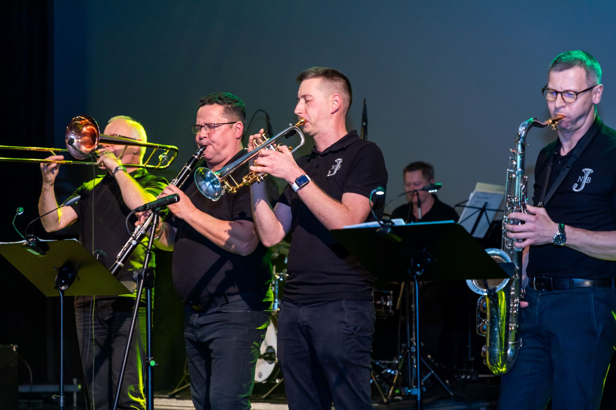 Na zdjęciu znajduje się zespół muzyczny grający na scenie. Widać czterech muzyków grających na różnych instrumentach dętych: puzonie, trąbce, saksofonie i trąbce. Muzycy są ubrani w czarne koszulki z logo 'NJB'. W tle widać perkusistę oraz stojaki na nuty. Scena jest oświetlona kolorowymi światłami, co dodaje dynamiki i atmosfery koncertowej.
