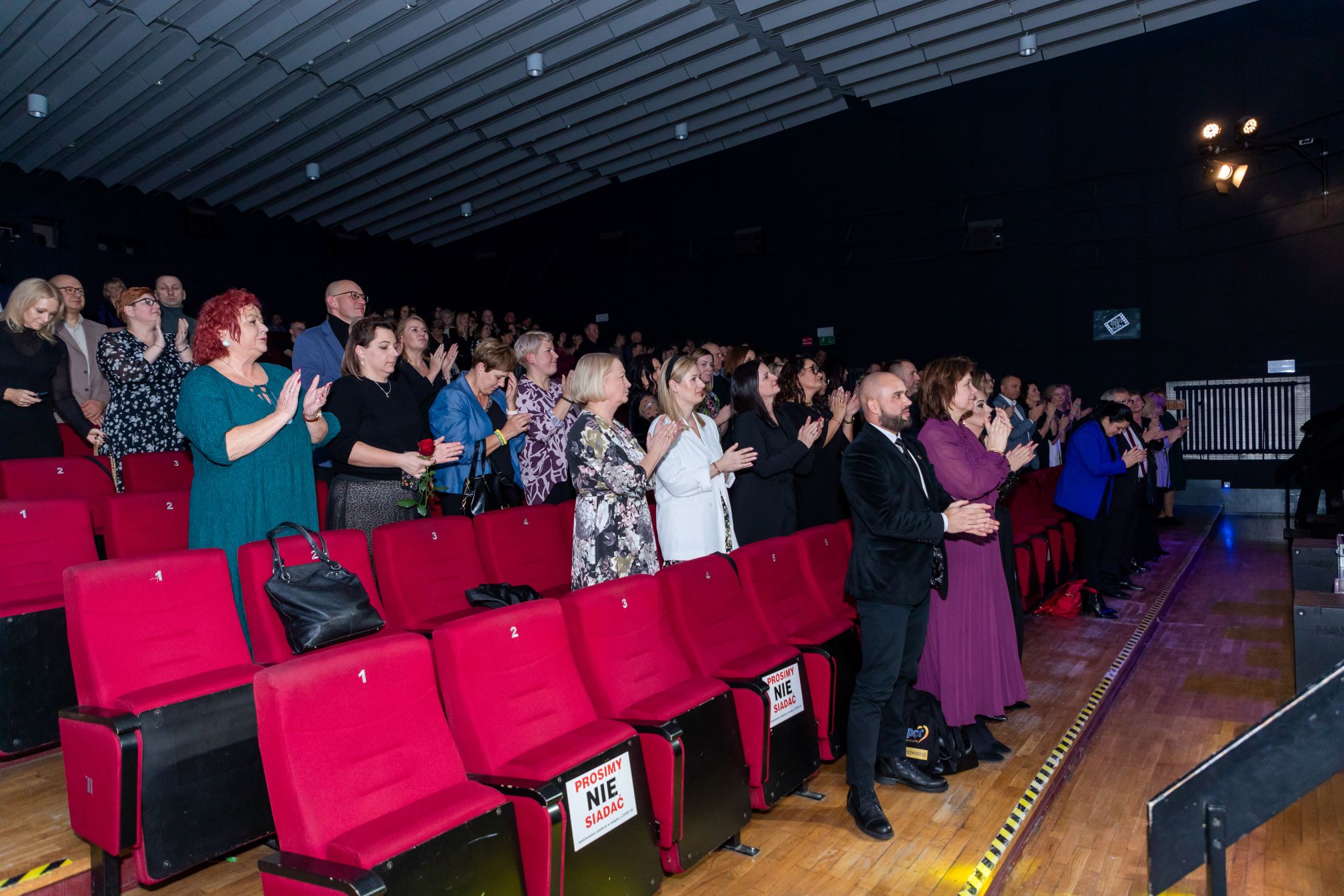 Na zdjęciu widać grupę ludzi stojących w sali kinowej. Wszyscy są ubrani elegancko i wydają się klaskać, co sugeruje, że właśnie zakończyło się jakieś przedstawienie lub występ. Czerwone fotele są ponumerowane, a na niektórych z nich znajdują się tabliczki z napisem 'PROSIMY NIE SIADAĆ'. W tle widać więcej osób, które również stoją i klaskają. Sala jest dobrze oświetlona, a sufit ma charakterystyczną, falistą strukturę.
