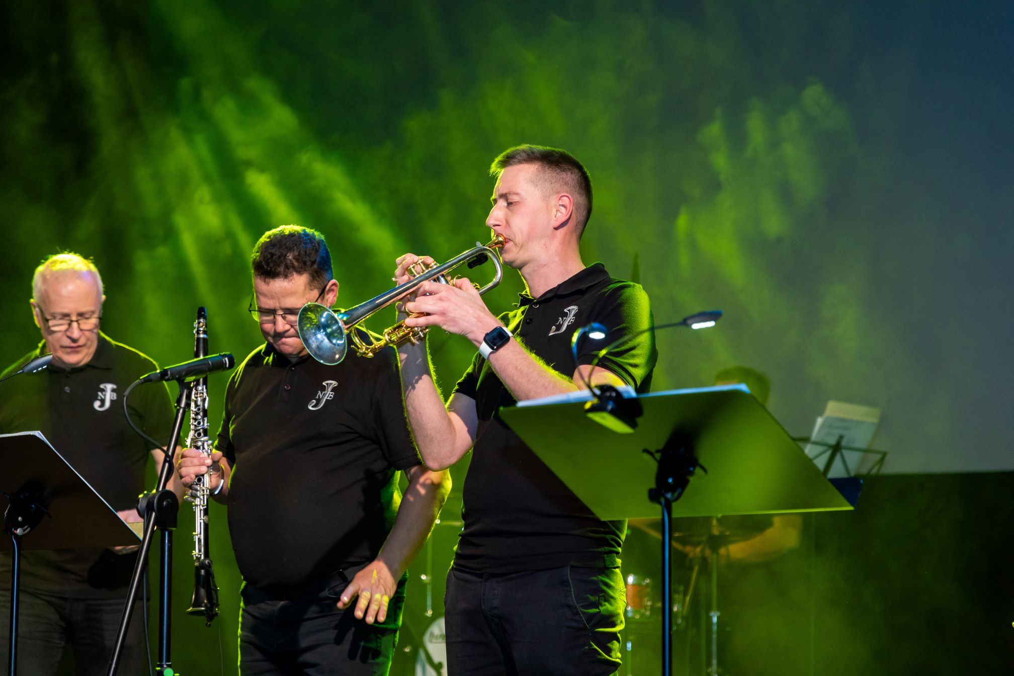 Na zdjęciu widać grupę muzyków występujących na scenie. Muzycy są ubrani w czarne koszulki z logo na lewej stronie klatki piersiowej. Jeden z muzyków gra na trąbce, inny na klarnecie, a w tle znajduje się trzeci muzyk. Scena jest oświetlona zielonymi światłami, co tworzy żywą atmosferę. Przed muzykami widoczne są stojaki na nuty z partyturami. Scena wydaje się być częścią koncertu lub występu na żywo.
