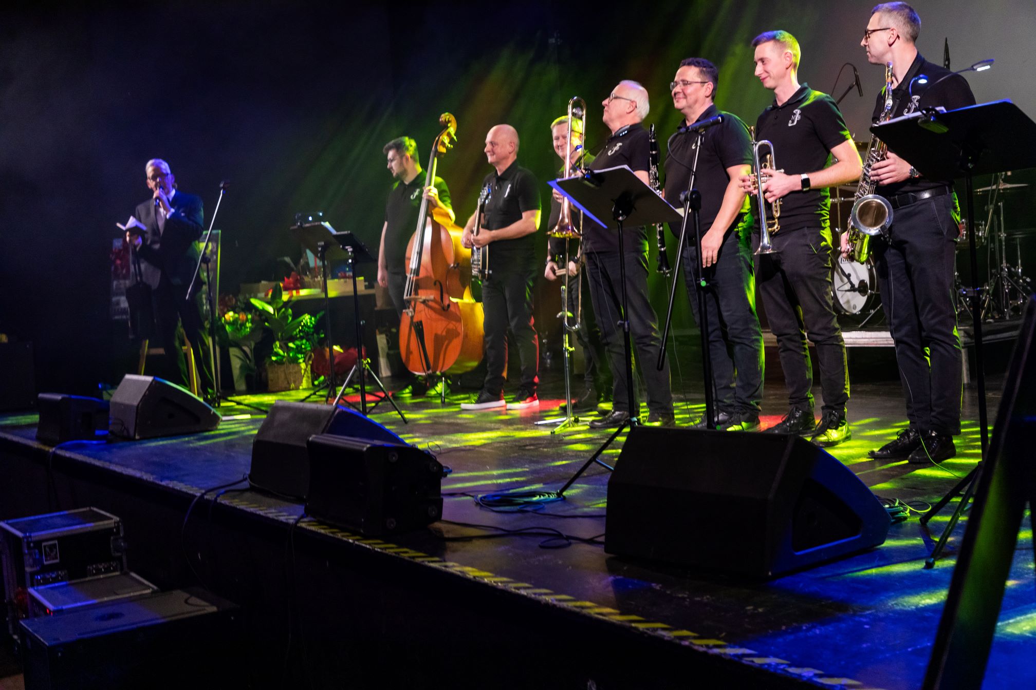 Na zdjęciu znajduje się zespół muzyczny występujący na scenie. W skład zespołu wchodzi kilku muzyków grających na różnych instrumentach, takich jak kontrabas, puzon, trąbka, saksofon i inne. Muzycy stoją w rzędzie, a przed nimi znajdują się mikrofony i pulpity na nuty. Scena jest oświetlona kolorowymi światłami, a w tle widać perkusję. Po lewej stronie sceny znajduje się osoba, która prawdopodobnie prowadzi wydarzenie lub zapowiada występ. Na scenie widoczne są również głośniki i inne elementy sprzętu nagłaśniającego.
