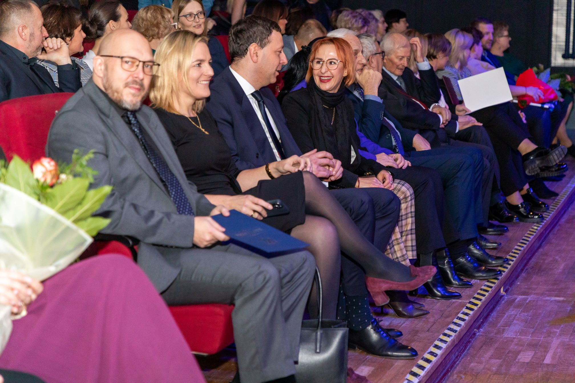 Zdjęcie przedstawia grupę ludzi siedzących w rzędzie, uczestniczących w formalnym wydarzeniu. Osoby są ubrane w eleganckie stroje, w tym garnitury i sukienki. Niektórzy z uczestników trzymają dokumenty lub teczki, a widoczne są również bukiety kwiatów. Miejsce przypomina audytorium lub teatr, z czerwonymi fotelami i drewnianą podłogą. Atmosfera sugeruje formalne zgromadzenie.
