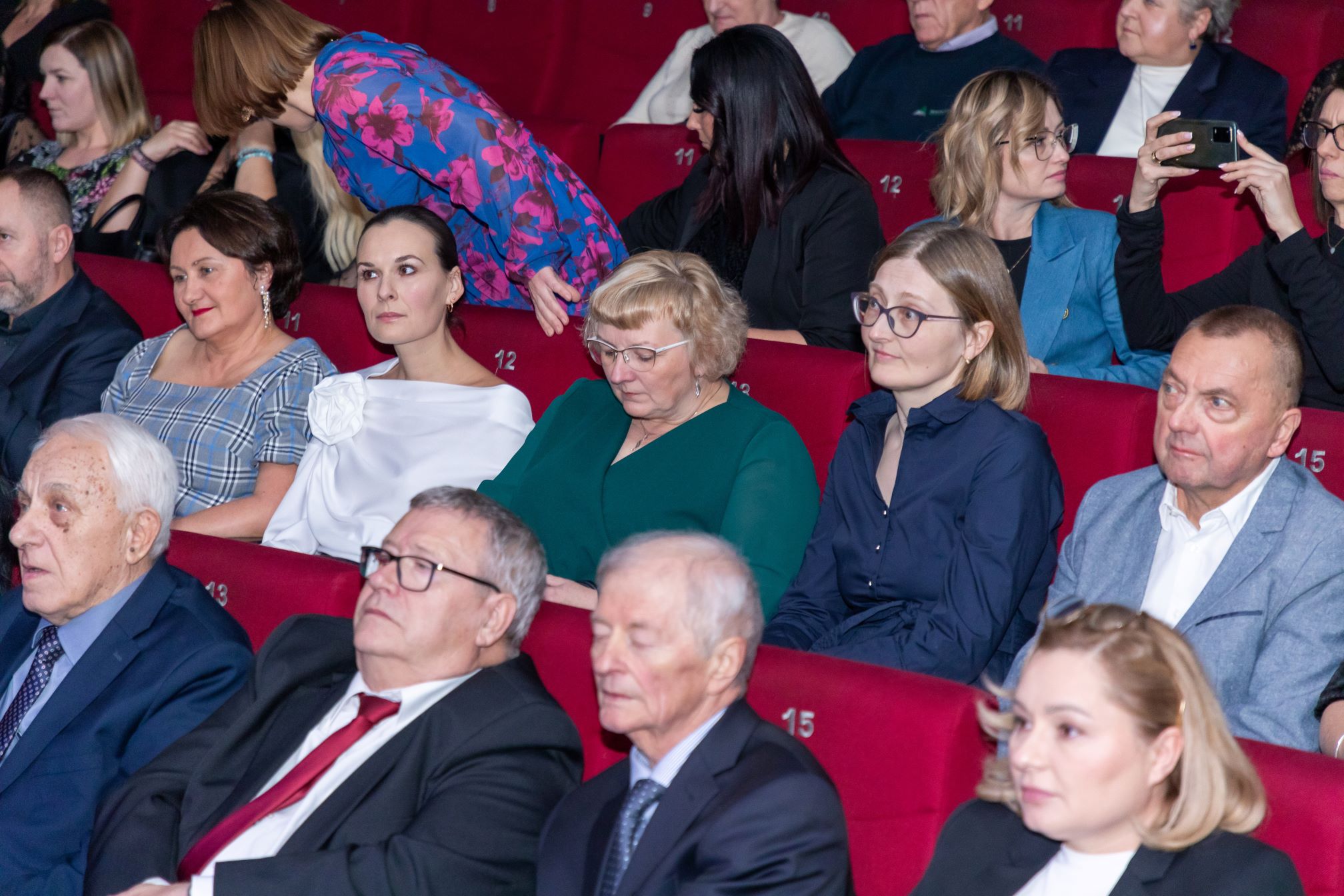 Zdjęcie przedstawia grupę ludzi siedzących w kinie. Wszyscy siedz ą na czerwonych fotelach, które są ponumerowane. Niektóre osoby są ubrane elegancko, w garnitury i sukienki, a inne mają na sobie bardziej casualowe ubrania. Jedna osoba w niebieskiej sukience z kwiatowym wzorem pochyla się, rozmawiając z kimś siedzącym. Inna osoba trzyma telefon i robi zdjęcie lub nagrywa. W tle widać więcej osób siedzących w rzędach.
