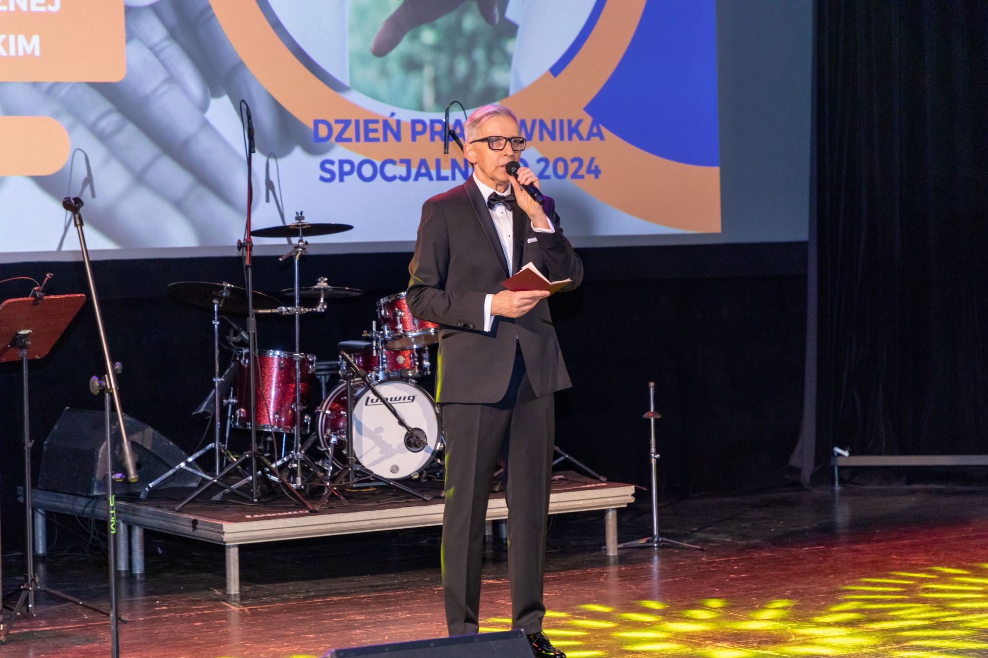 Na zdjęciu znajduje się mężczyzna w garniturze, który stoi na scenie i trzyma mikrofon oraz notes. W tle widać zestaw perkusyjny marki Ludwig oraz ekran z napisem 'DZIEŃ PRACOWNIKA SOCJALNEGO 2024'. Scena jest oświetlona reflektorami, a na podłodze widać kolorowe światła.
