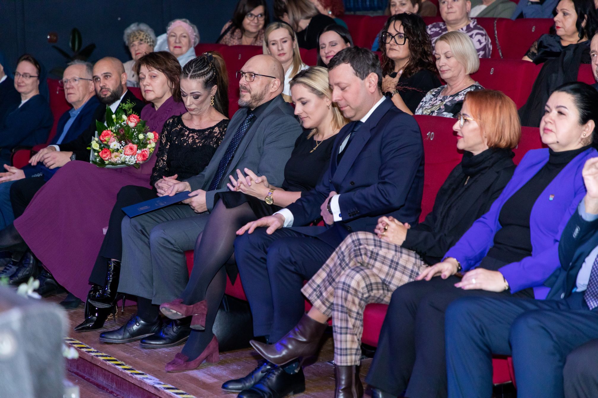 Na zdjęciu widać grupę ludzi siedzących w kinie. Osoby są elegancko ubrane, niektóre trzymają bukiety kwiatów. Wygląda na to, że uczestniczą w formalnym wydarzeniu, takim jak występ lub prezentacja. Widzowie wydają się być zaangażowani i uważnie obserwują przebieg wydarzeń, co podkreśla wagę i znaczenie tego wydarzenia.
