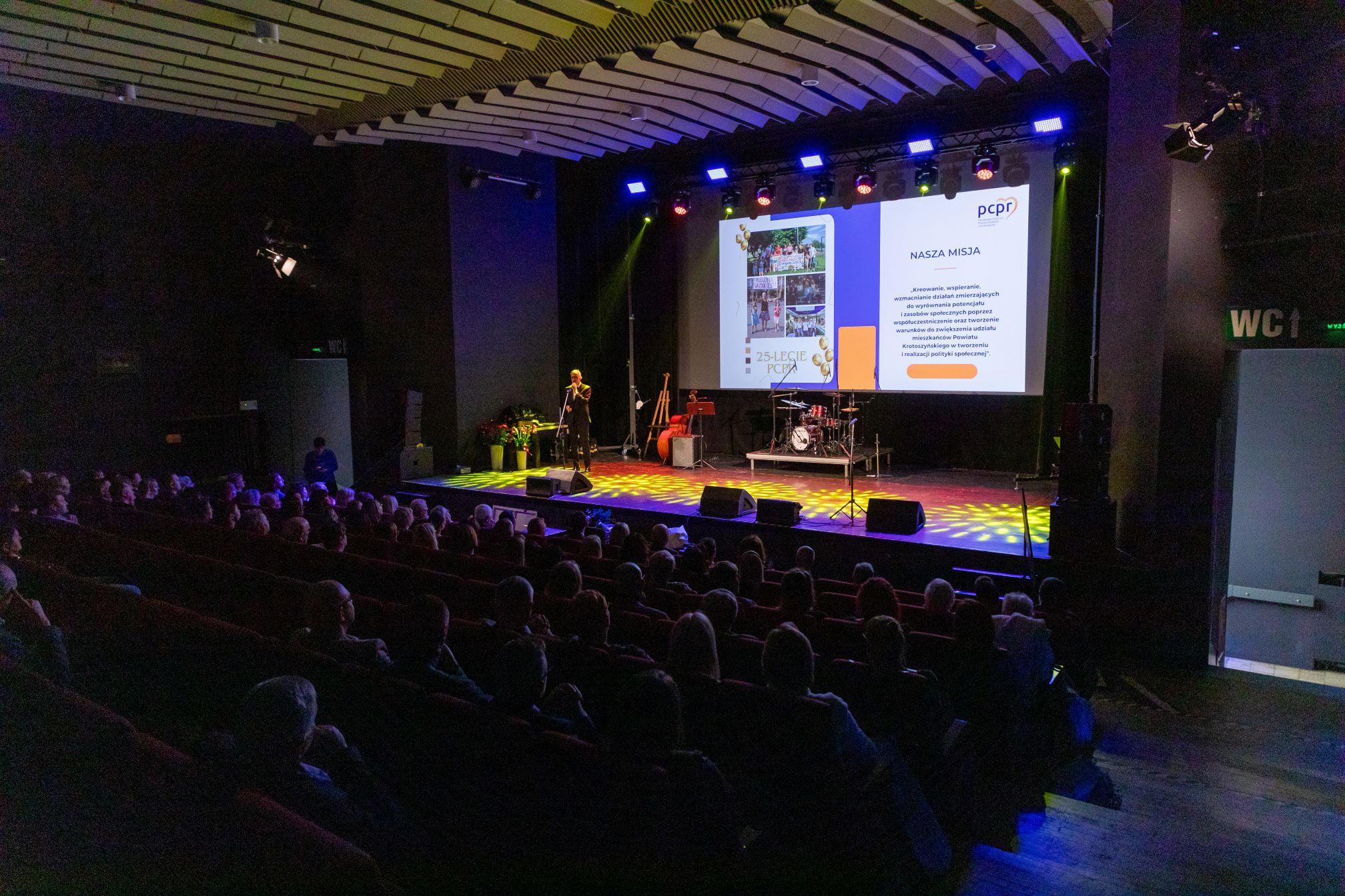 Na zdjęciu znajduje się scena w sali kinowej. Na scenie stoi osoba przemawiająca do publiczności. W tle znajduje się duży ekran, na którym wyświetlana jest prezentacja. Na ekranie widnieje tekst 'NASZA MISJA' oraz kilka zdjęć i logo 'PCPR'. Na scenie znajdują się również instrumenty muzyczne, takie jak perkusja i kontrabas, co sugeruje, że może odbywać się tam również występ muzyczny. Publiczność siedzi w rzędach przed sceną, a sala jest oświetlona różnokolorowymi światłami.
