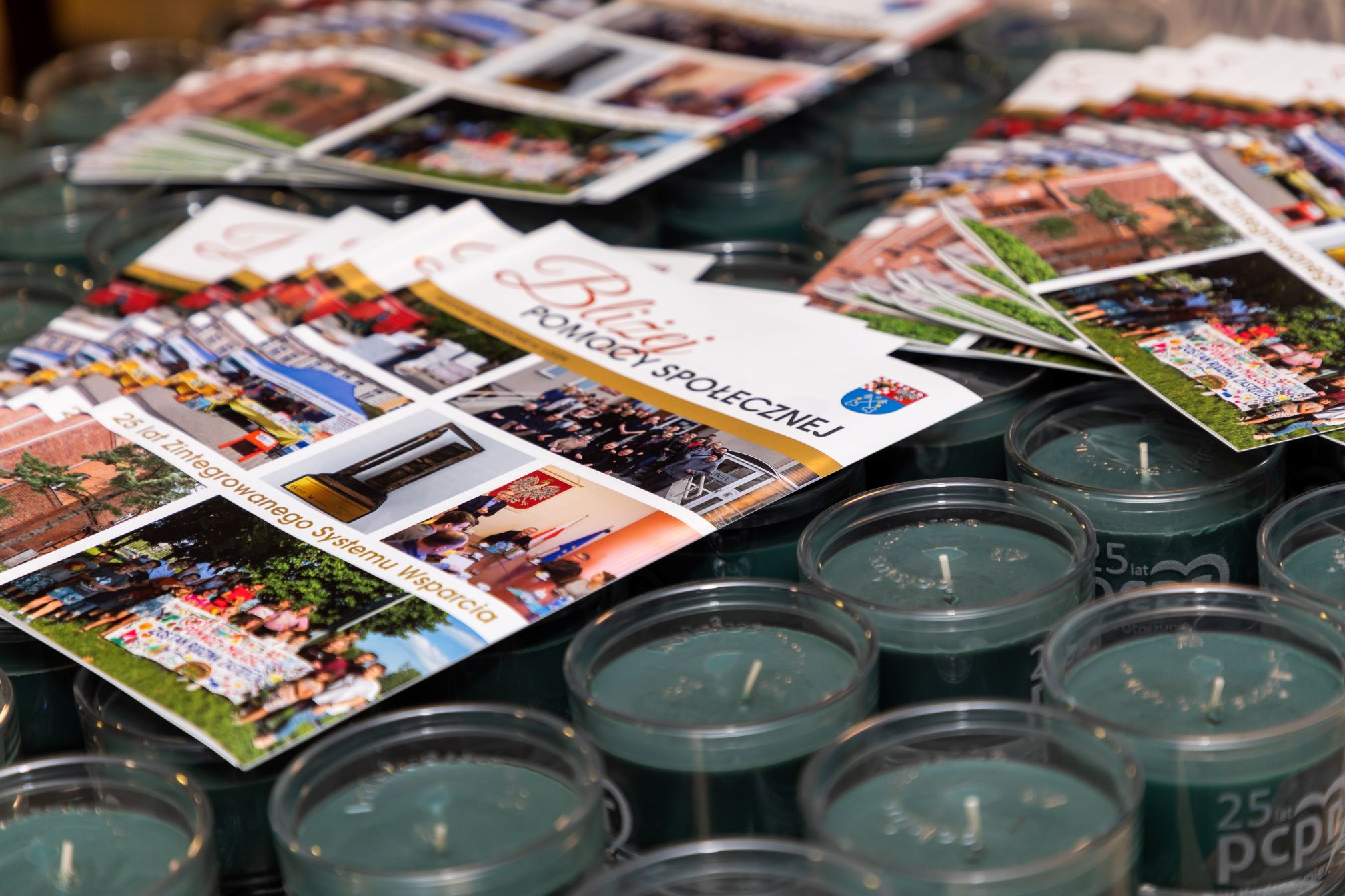 Na zdjęciu widoczne są biuletyny leżące na świecach. Mają tytuł 'Bliżej POMOCY SPOŁECZNEJ' i zawierają kilka zdjęć przedstawiających różne wydarzenia i osoby. Na biuletynach znajduje się również herb oraz napis '25 lat Zintegrowanego Systemu Wsparcia'. Świece są koloru zielonego i mają napis '25 lat PCPR
