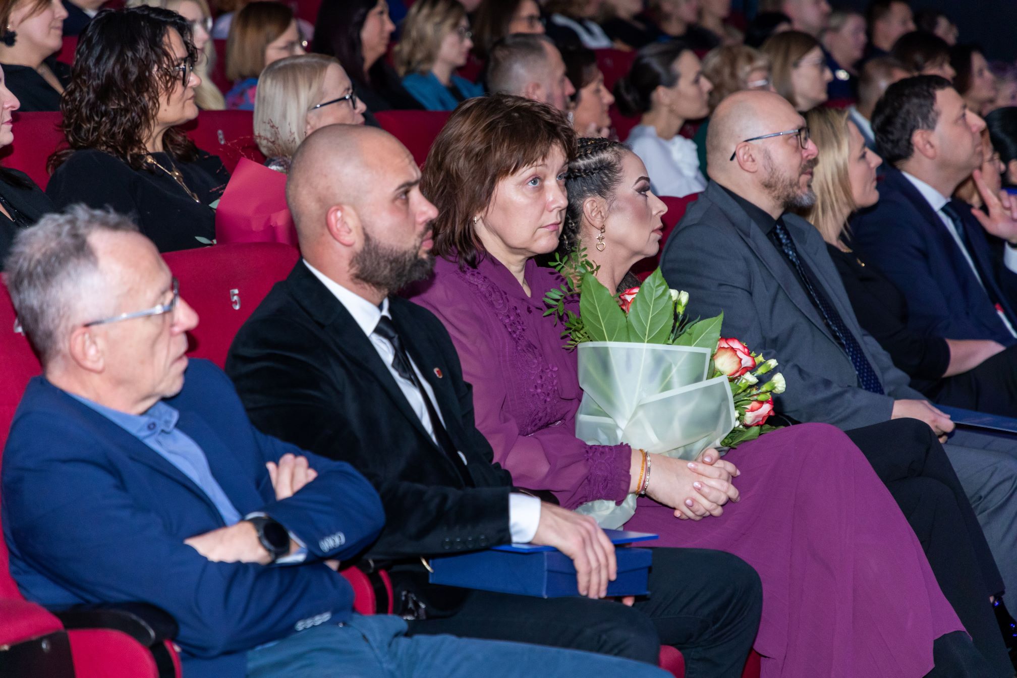 Na zdjęciu widać grupę ludzi siedzących w rzędach czerwonych foteli w sali kinowej. Osoby są elegancko ubrane, co sugeruje, że uczestniczą w jakimś oficjalnym wydarzeniu, takim jak gala, ceremonia wręczenia nagród lub uroczystość. Jedna z osób trzyma bukiet kwiatów, co może wskazywać na to, że jest to osoba wyróżniona lub nagrodzona. W tle widać więcej osób siedzących w podobnych rzędach.
