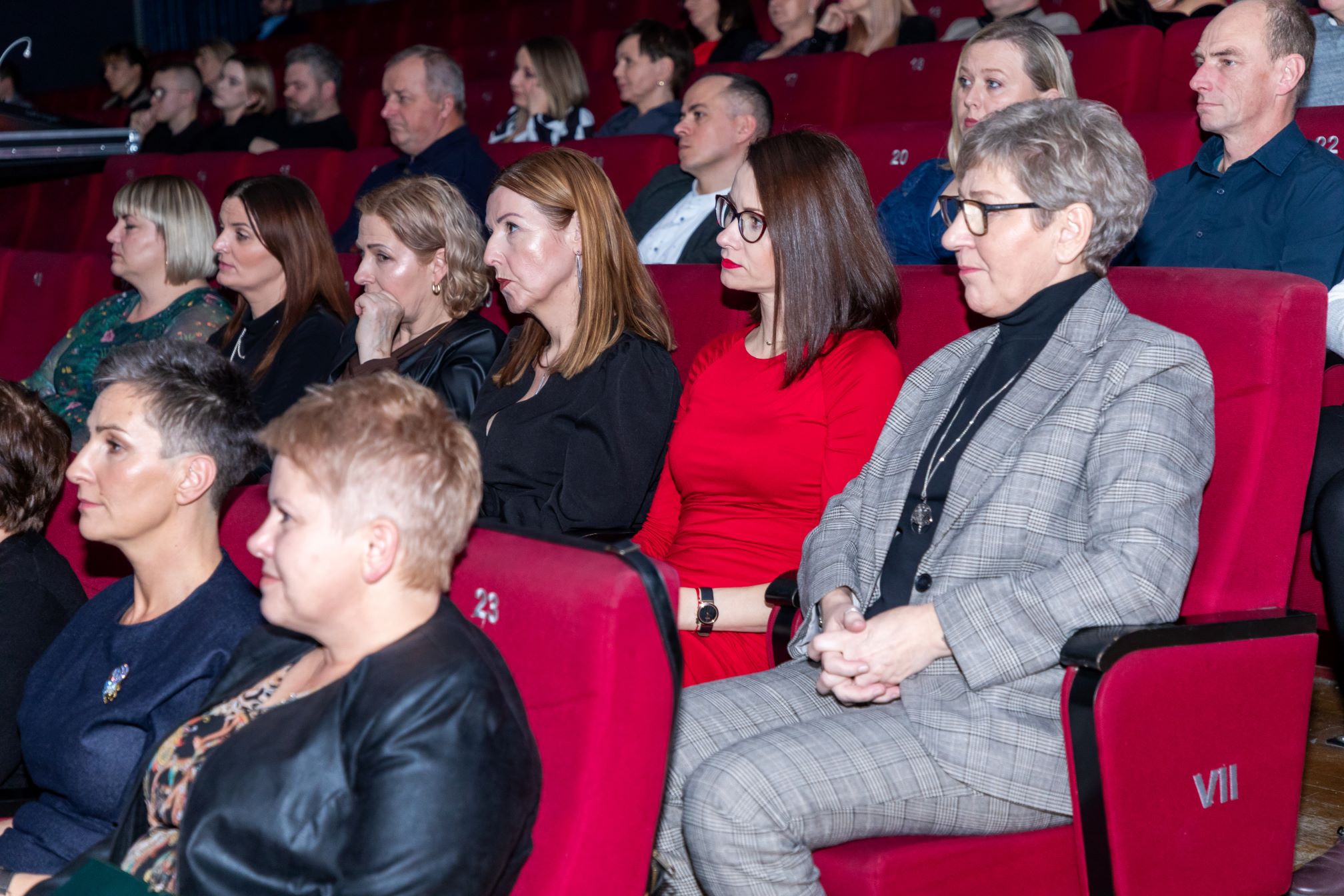 Zdjęcie przedstawia grupę ludzi siedzących w kinie. Wszyscy siedzą na czerwonych fotelach, które są ponumerowane. Osoby na zdjęciu są ubrane w eleganckie stroje, co sugeruje, że uczestniczą w jakimś formalnym wydarzeniu. Na pierwszym planie widać kobietę w czerwonej sukience oraz kobietę w szarym garniturze w kratkę. W tle widać więcej osób siedzących w rzędach za nimi.
