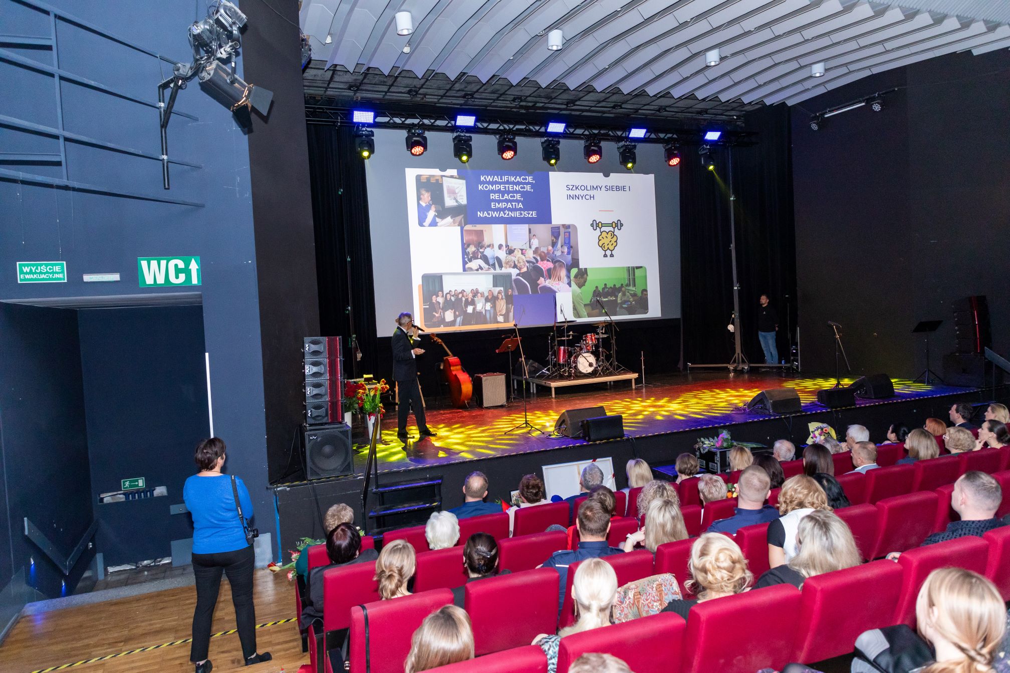 Zdjęcie przedstawia salę kinową, w której odbywa się prezentacja. Na pierwszym planie widać publiczność siedzącą na czerwonych fotelach, skierowaną w stronę sceny. Na scenie znajduje się osoba prowadząca prezentację, a za nią na dużym ekranie wyświetlane są slajdy. Na slajdach widoczne są zdjęcia grup ludzi oraz tekst w języku polskim: 'KWALIFIKACJE, KOMPETENCJE, RELACJE, EMPATIA NAJWAŻNIEJSZE' oraz 'SZKOLIMY SIEBIE I INNYCH'. Na scenie znajdują się również instrumenty muzyczne, takie jak perkusja i kontrabas.
