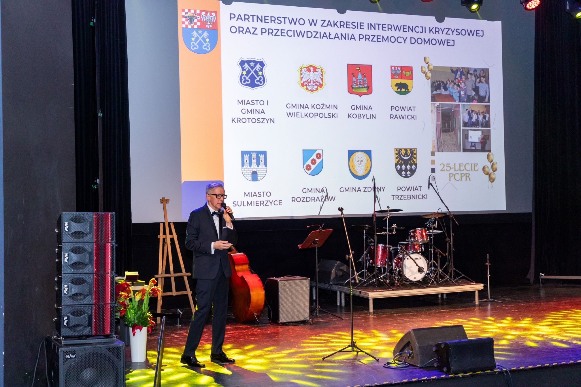 Na zdjęciu znajduje się mężczyzna w garniturze, który przemawia na scenie. W tle widoczny jest duży ekran z herbami różnych miast i gmin oraz napisem 'PARTNERSTWO W ZAKRESIE INTERWENCJI KRYZYSOWEJ ORAZ PRZECIWDZIAŁANIA PRZEMOCY DOMOWEJ'. Na ekranie znajdują się herby oraz nazwy miast i gmin: Miasto i Gmina Krotoszyn, Gmina Koźmin Wielkopolski, Gmina Kobylin, Powiat Rawicki, Miasto Sulmierzyce, Gmina Rozdrażew, Gmina Zduny, Powiat Trzebnicki. Po prawej stronie ekranu znajduje się logo z napisem '25-LECIE PCPR'. Na scenie widoczne są również instrumenty muzyczne, takie jak perkusja i kontrabas, oraz mikrofony i głośniki.
