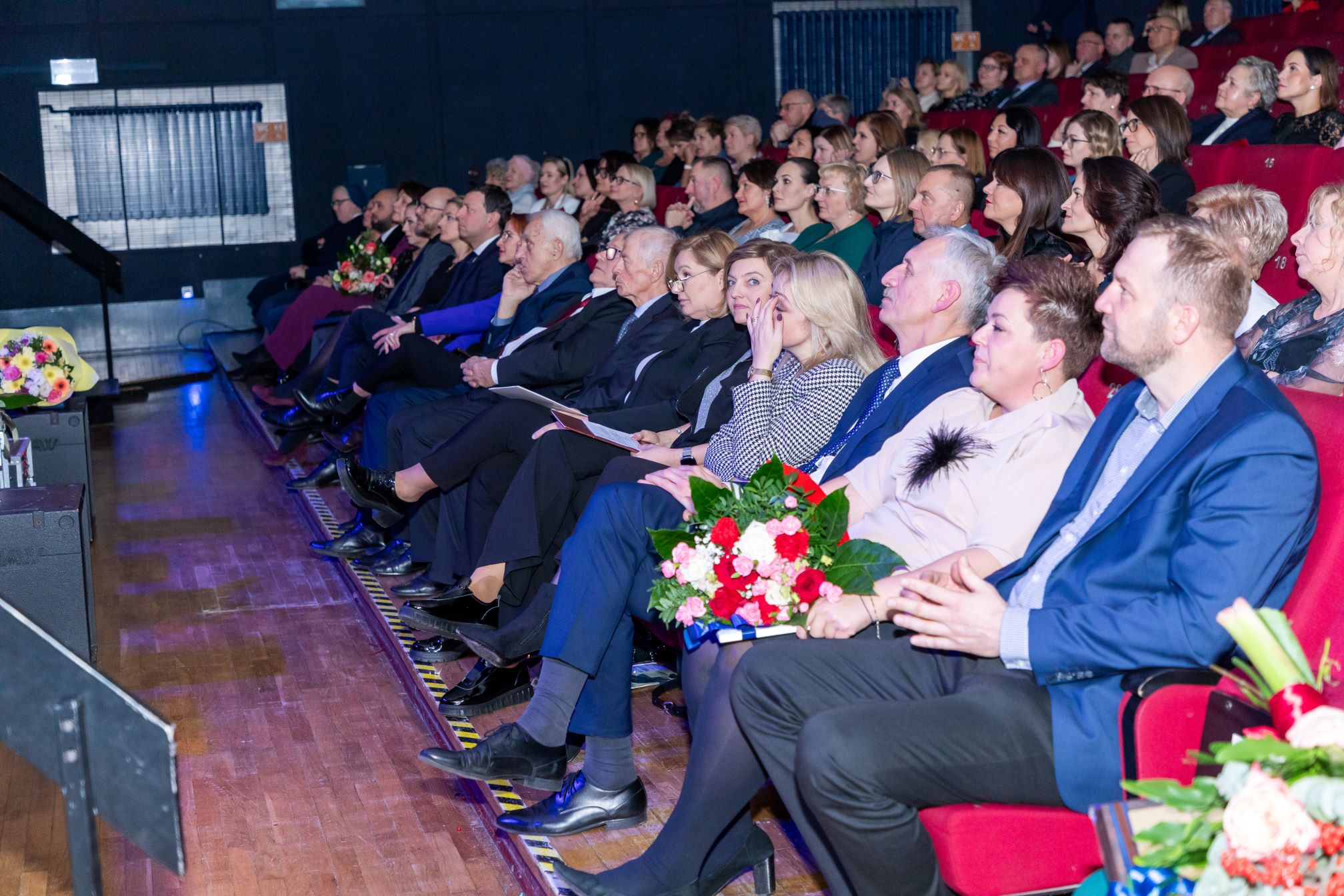 Zdjęcie przedstawia grupę ludzi siedzących w rzędach na widowni w sali kinowej. Wszyscy są elegancko ubrani, niektórzy trzymają bukiety kwiatów. Na pierwszym planie widać osoby siedzące w pierwszym rzędzie, a w tle kolejne rzędy widowni. Sala jest dobrze oświetlona, a na podłodze widać drewniany parkiet. Na scenie po lewej stronie znajduje się podium lub mównica.
