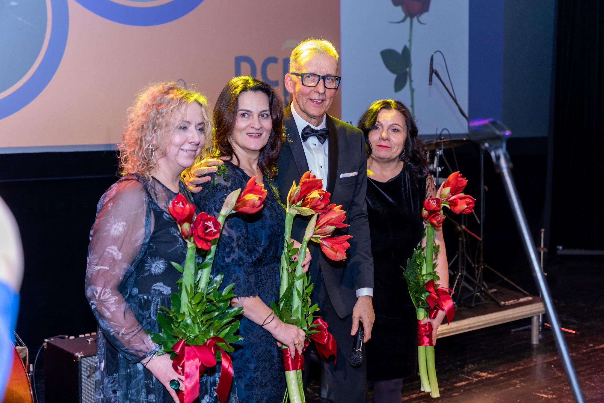 Na zdjęciu znajduje się czterech ludzi stojących na scenie. Trzy osoby trzymają bukiety czerwonych kwiatów z zielonymi liśćmi, przewiązane czerwonymi wstążkami. Jedna z osób, ubrana w garnitur, trzyma mikrofon. W tle widać fragmenty dekoracji scenicznej, w tym duży plakat z różą oraz fragmenty instrumentów muzycznych.
