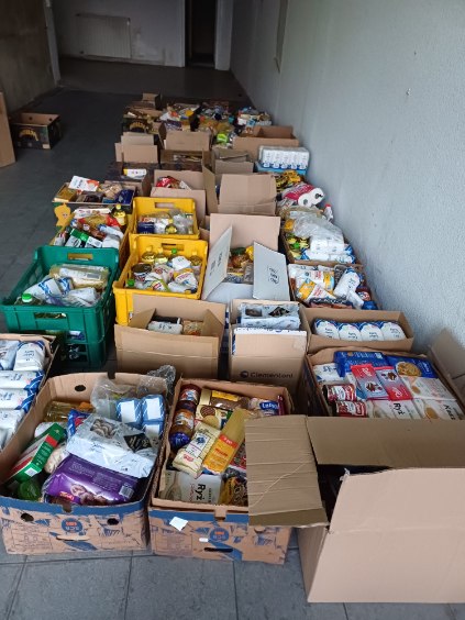 Zdjęcie przedstawia rząd pudełek i skrzynek wypełnionych różnymi produktami spożywczymi i artykułami codziennego użytku. W pudełkach znajdują się między innymi makarony, konserwy, płatki śniadaniowe, mleko, papier toaletowy, olej, cukier, mąka oraz inne produkty. Pudełka są ustawione wzdłuż ściany w pomieszczeniu