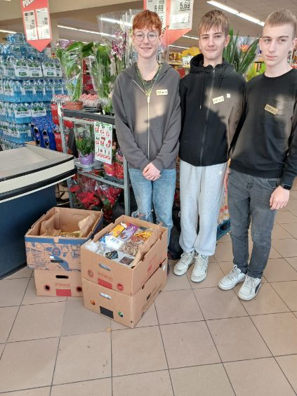 Zdjęcie przedstawia trzy osoby stojące w sklepie w pobliżu ekspozycji kwiatów i wody butelkowanej. Osoby te stoją obok dwóch kartonowych pudeł wypełnionych różnymi przedmiotami. Ubrane są w codzienne ubrania i mają plakietki z imionami przypięte do koszulek