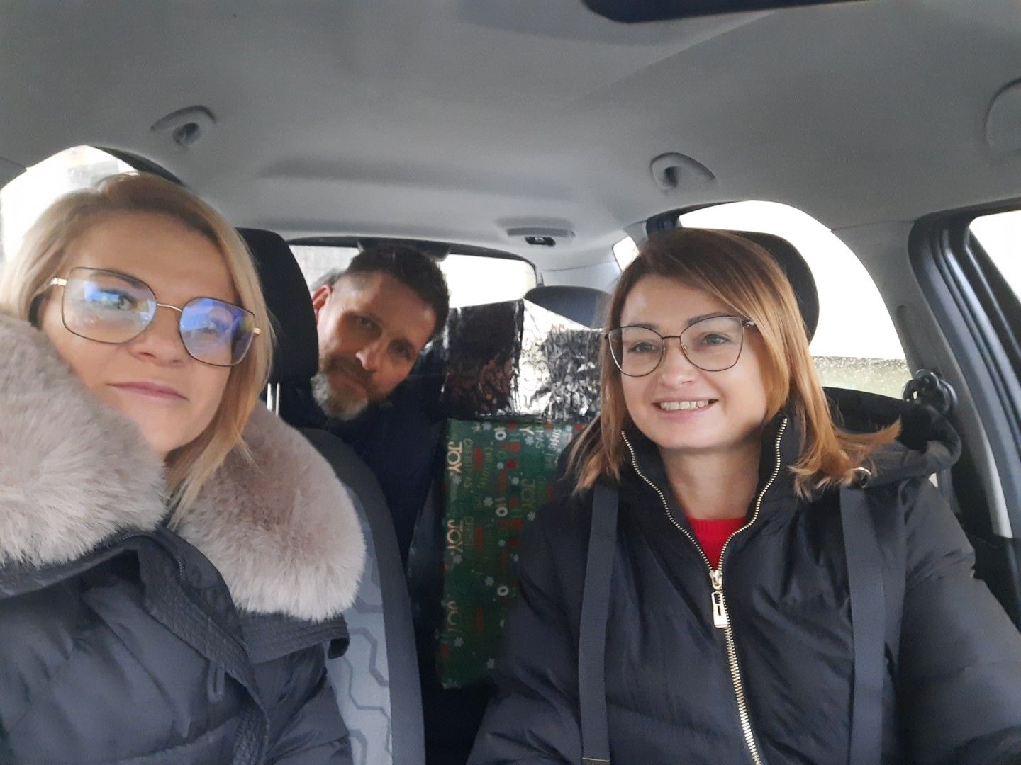 Wnętrze samochodu z trzema osobami: dwie siedzą z przodu, jedna z tyłu. Przednie osoby mają na sobie zimowe kurtki; osoba po lewej nosi okulary. Na tylnym siedzeniu widać duże, zapakowane prezenty.
