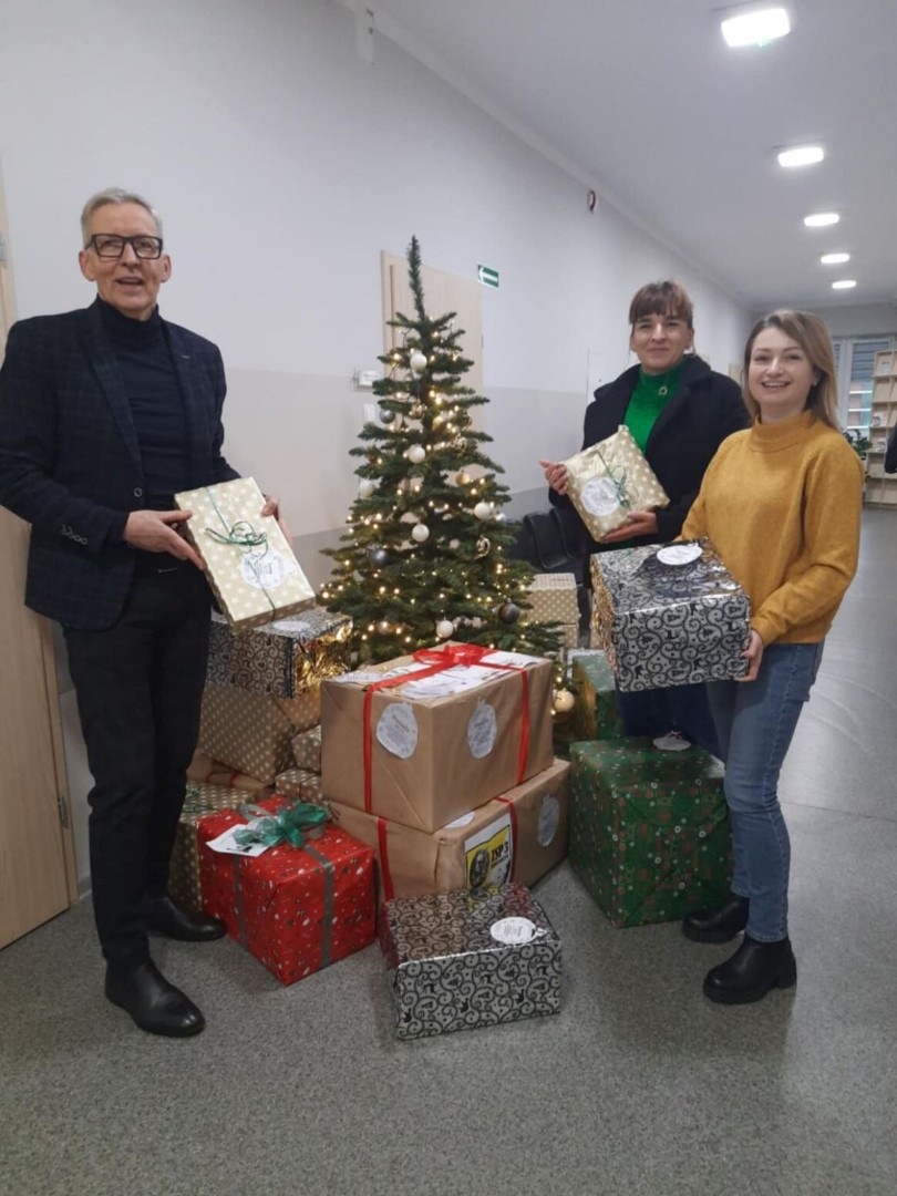 Scena na korytarzu. Trzy osoby stoją wokół małej udekorowanej choinki. Trzymają zapakowane prezenty, a kilka innych znajduje się pod drzewkiem. Jest to świąteczna zbiórka prezentów. Choinka jest udekorowana światełkami i ozdobami, co dodaje świątecznego nastroju. Zapakowane prezenty są w różnych rozmiarach i kolorach, co wskazuje na różnorodność upominków.
